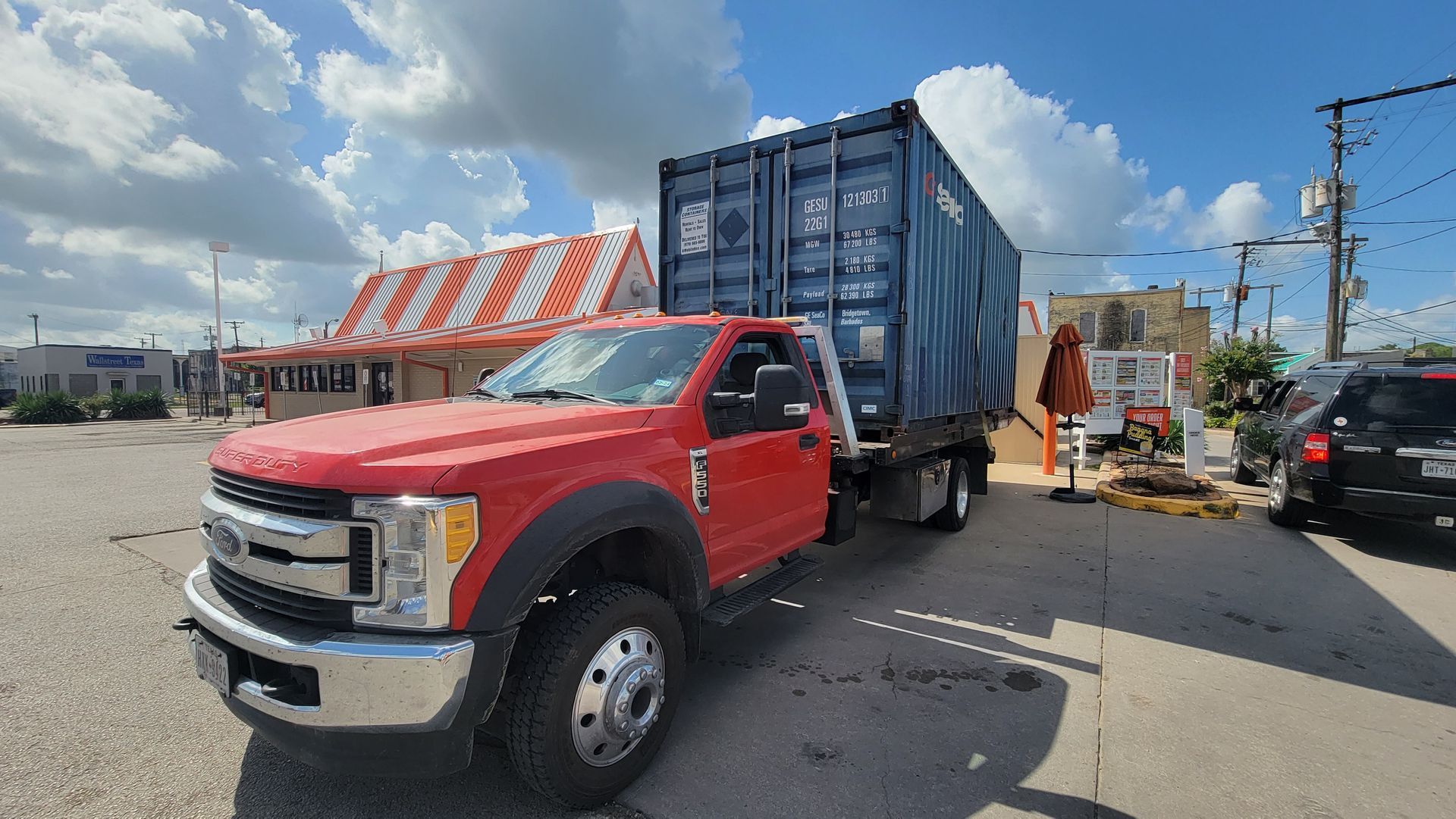 Red dump truck hauling a blue container on a street near construction barriers and a worker cart