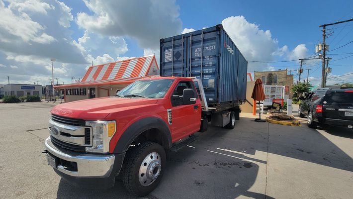Red semi-truck hauling a blue shipping container on a street near orange barriers and a black car