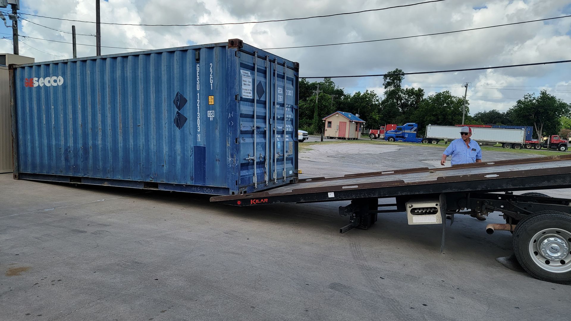 Blue shipping container on a flatbed trailer in a parking lot, with trees and a cloudy sky behind.