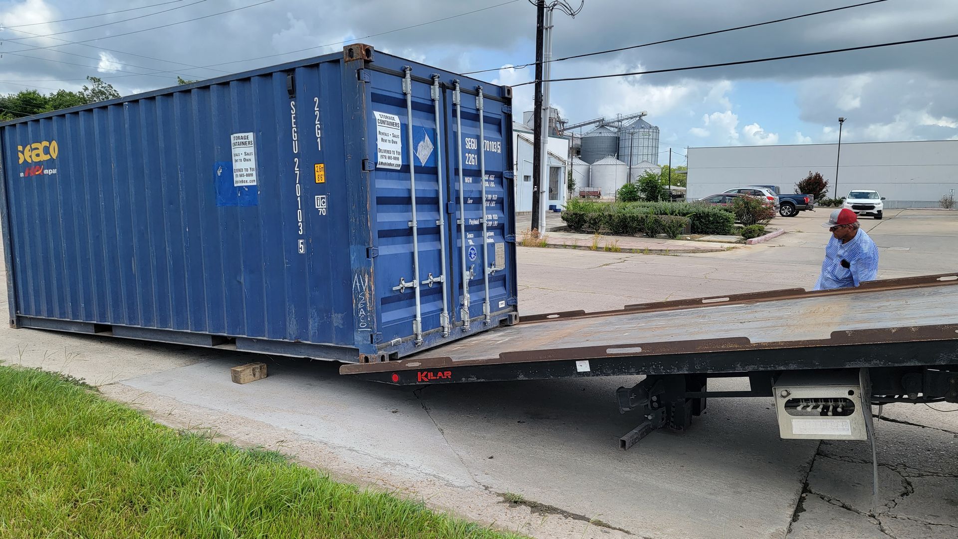 Blue shipping container on a flatbed trailer in an industrial area, with a worker nearby in blue and red clothes
