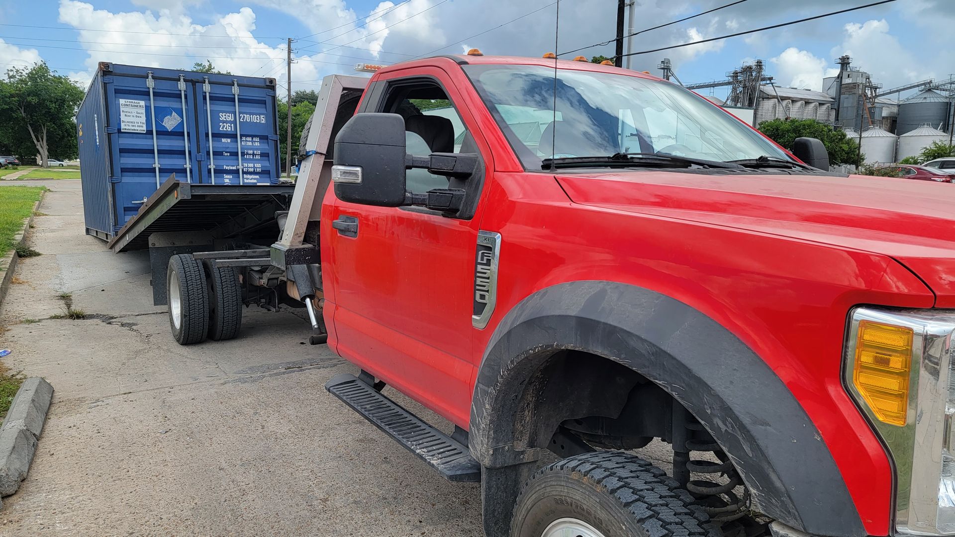 Red pickup truck towing a blue trailer on a roadside, viewed from the front side