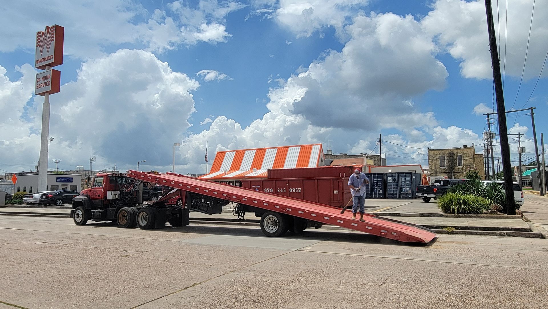 Tow truck hauling a car up a red ramp on a street near an orange-striped tent under a cloudy sky
