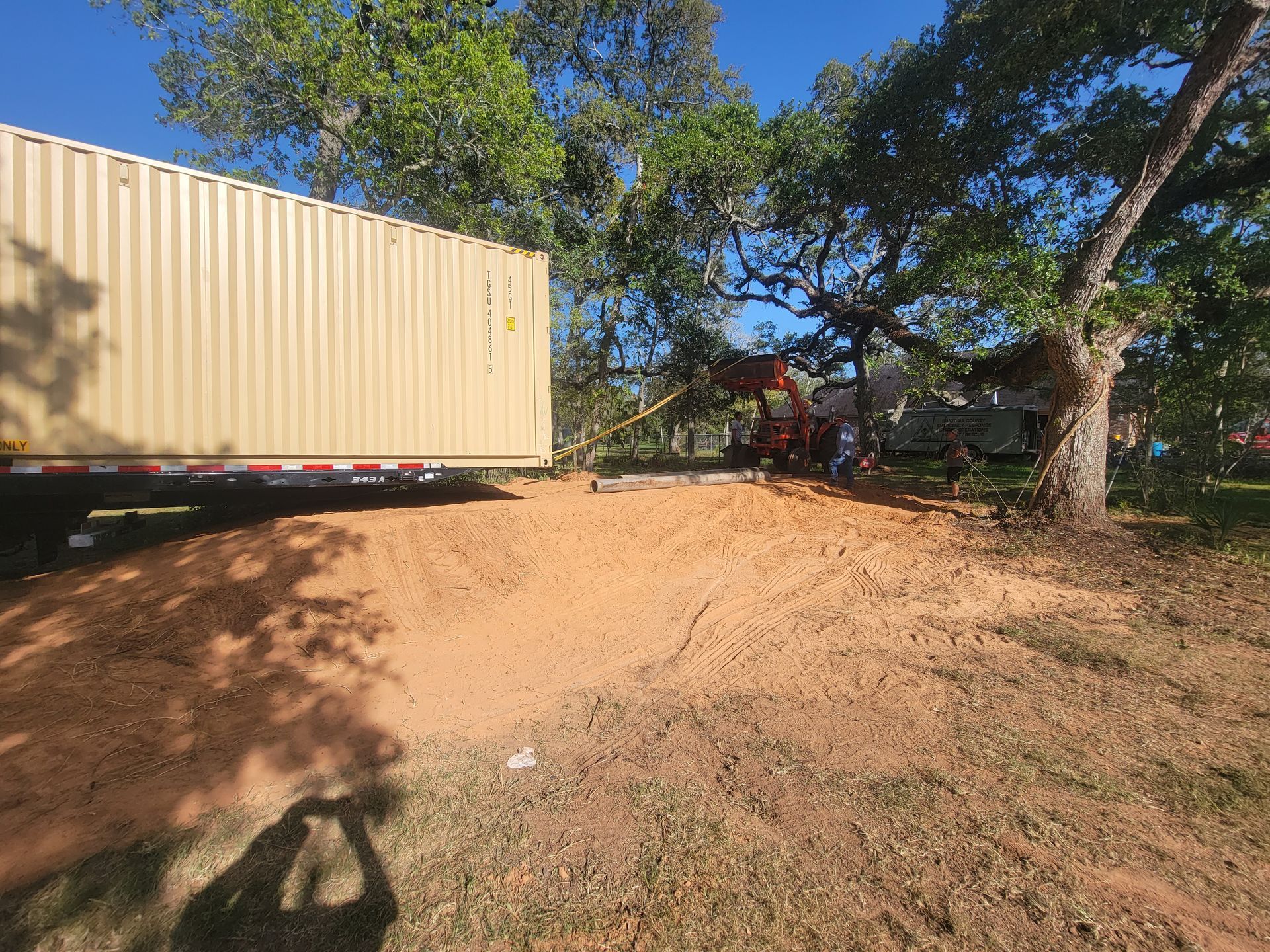 Yellow shipping container beside a dirt lot with trees and a red tractor in the background