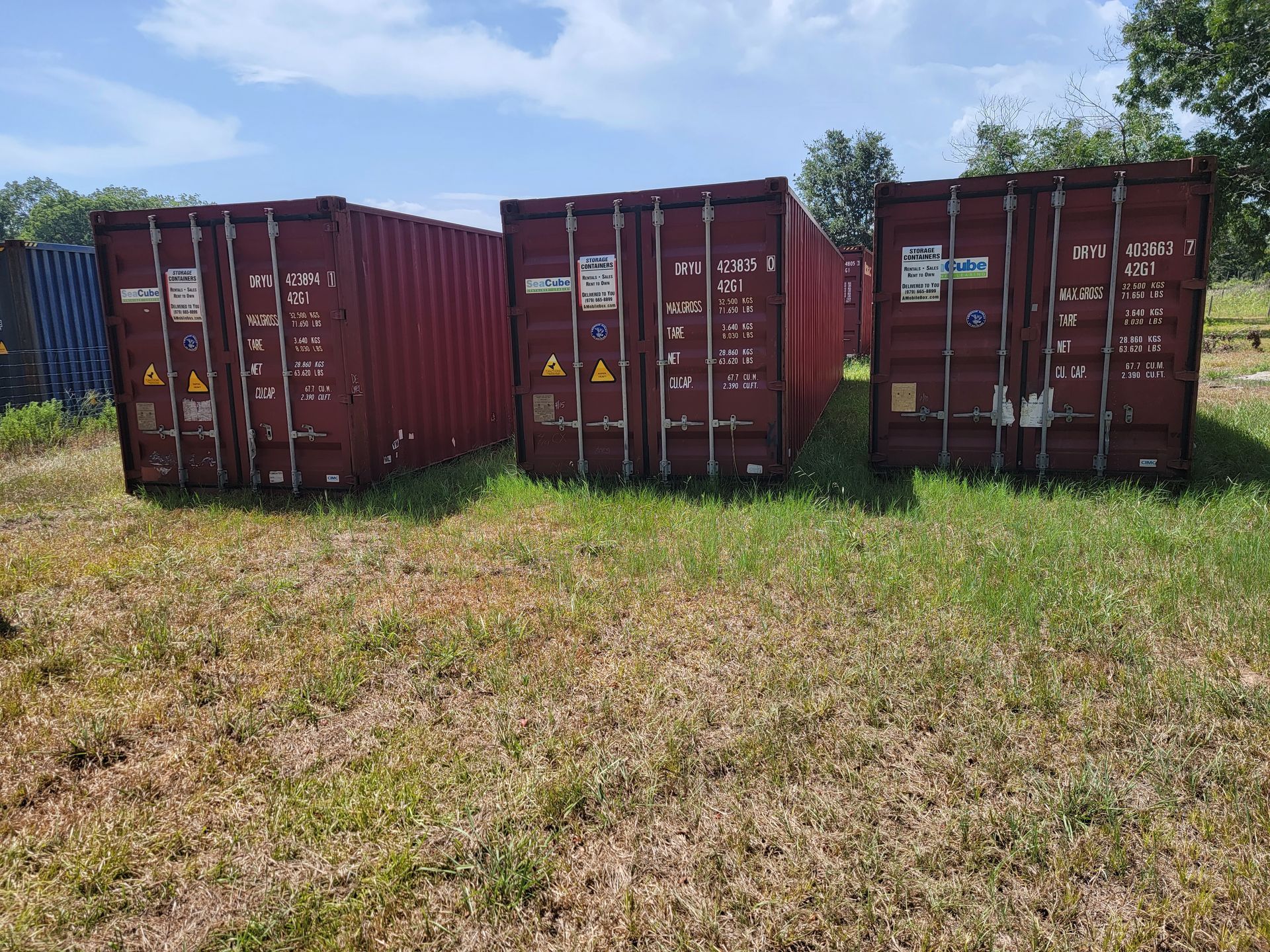 Three rust-red shipping containers on grass under a partly cloudy sky.