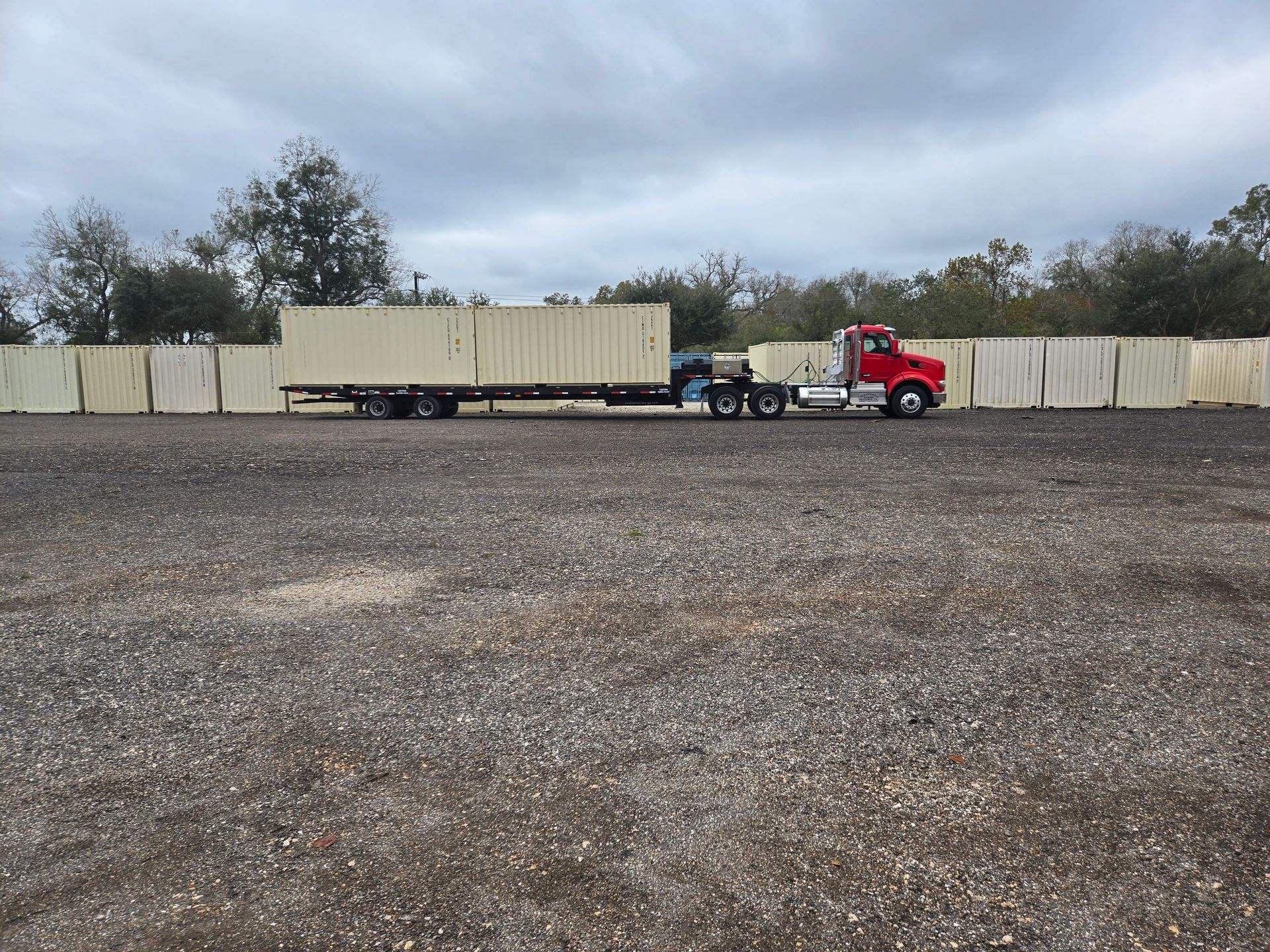 Red semi-truck with beige trailer parked on a gravel lot under cloudy sky