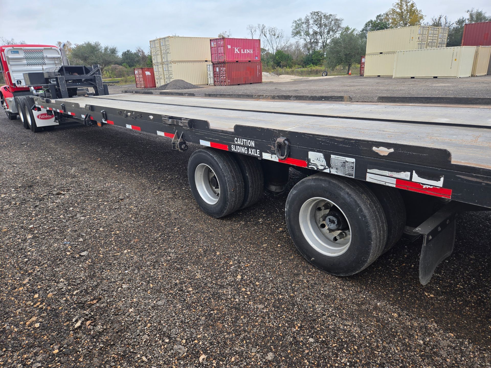 Flatbed trailer parked on gravel lot, with dual wheels and red reflectors under an overcast sky