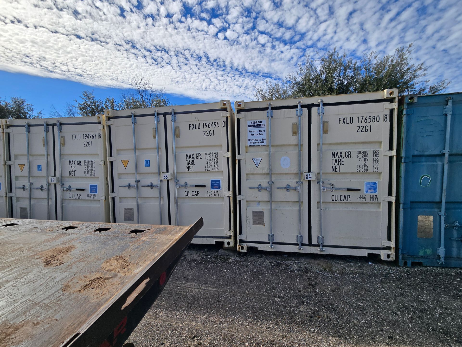 Row of white and blue shipping containers on gravel under a cloudy sky
