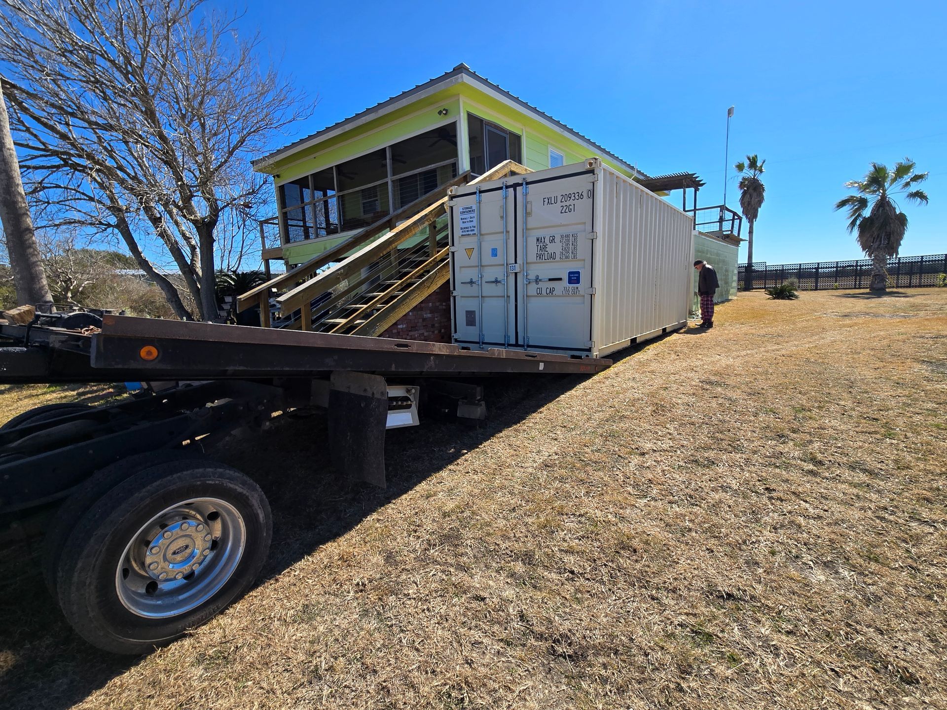 Flatbed truck carrying a white shipping container outside a lime-green house on a dry lot