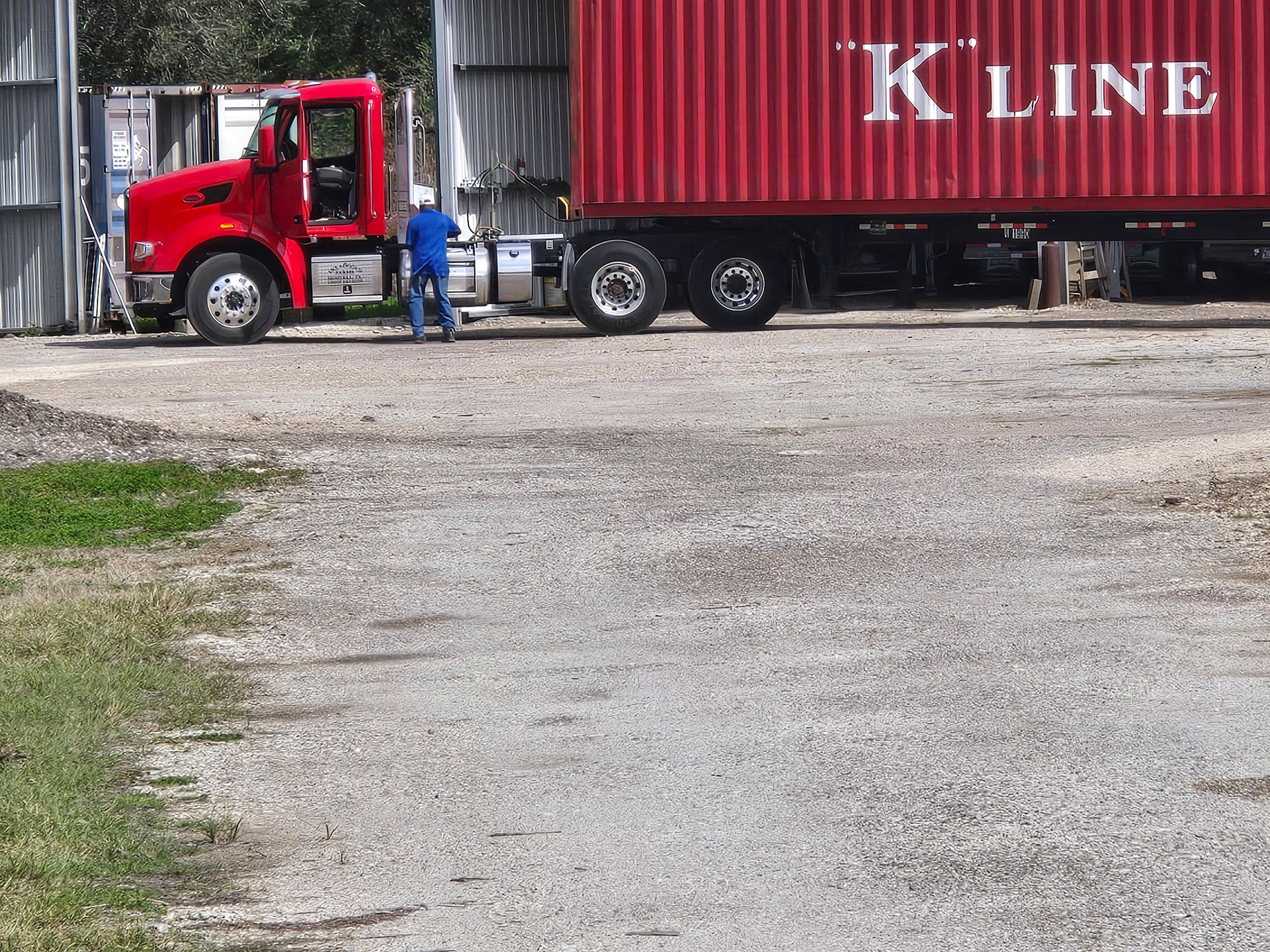 Red semi-truck parked beside a red K LINE shipping container, with a person standing nearby on gravel.