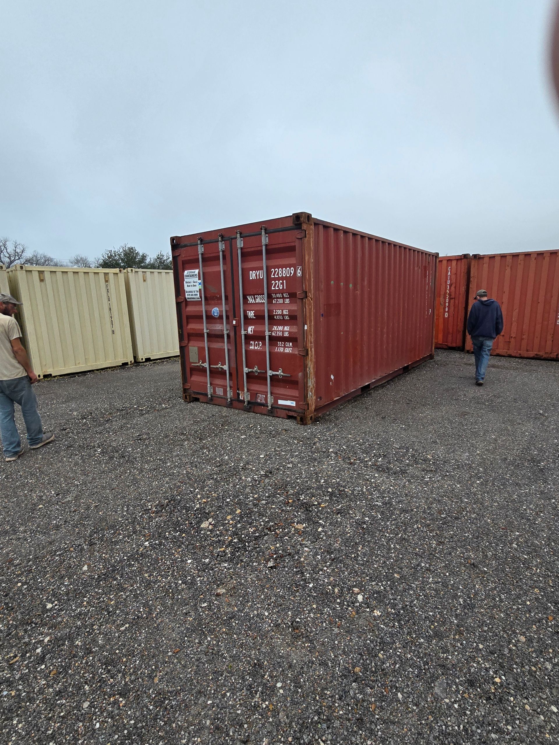 Red shipping container on gravel lot with two people nearby and a beige container in the background