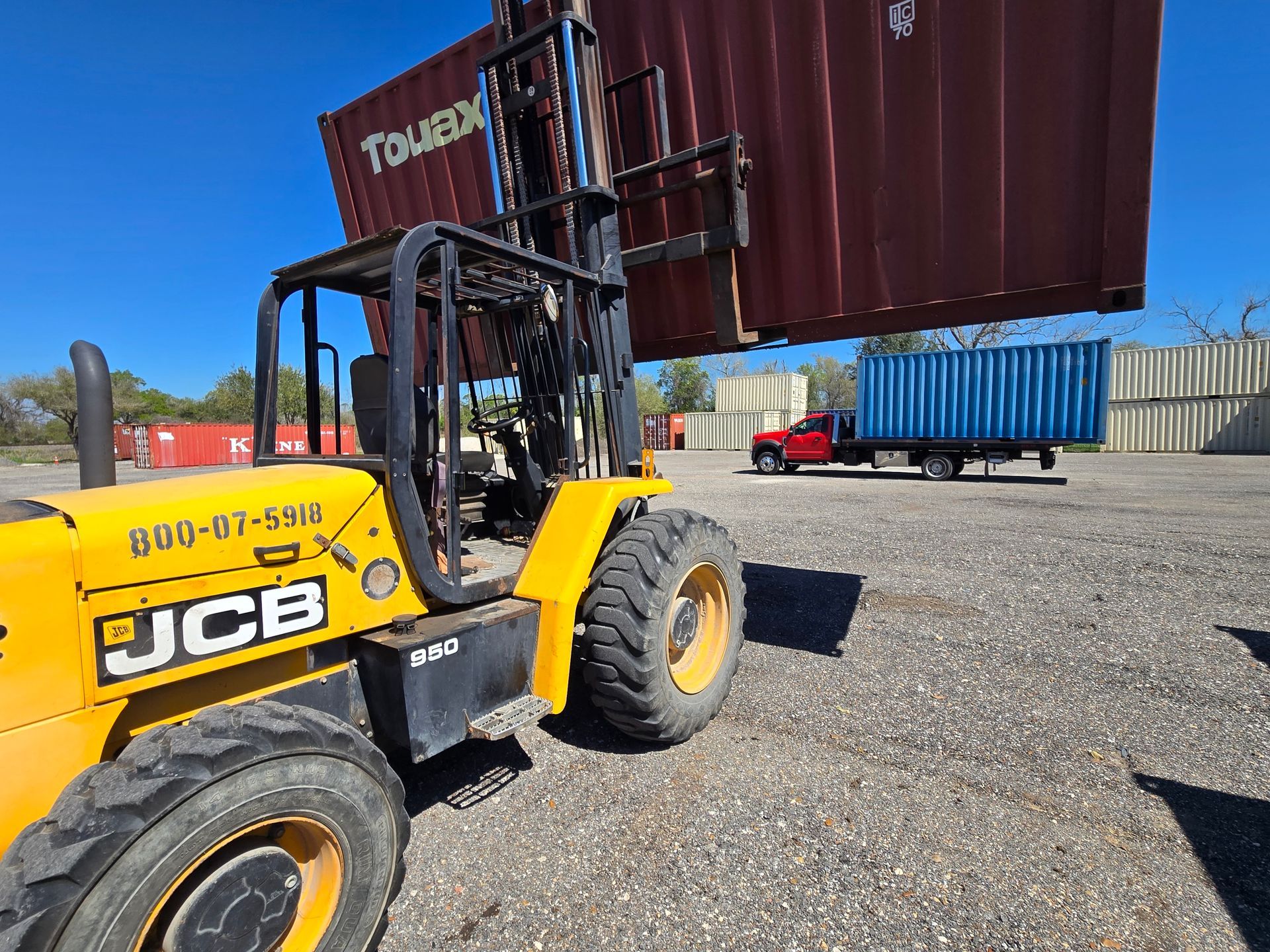Yellow JCB forklift lifting a red shipping container in a gravel yard