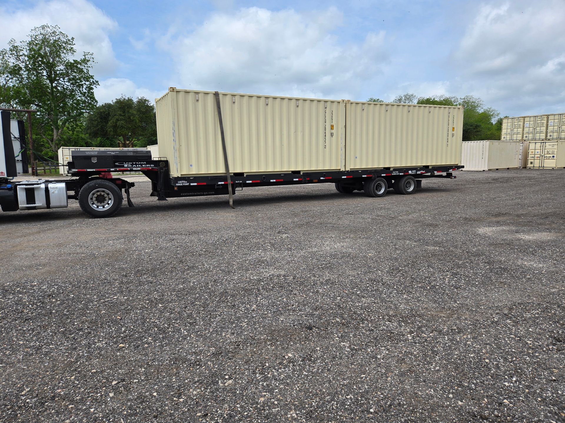 Flatbed truck hauling a beige shipping container in a gravel lot under a partly cloudy sky