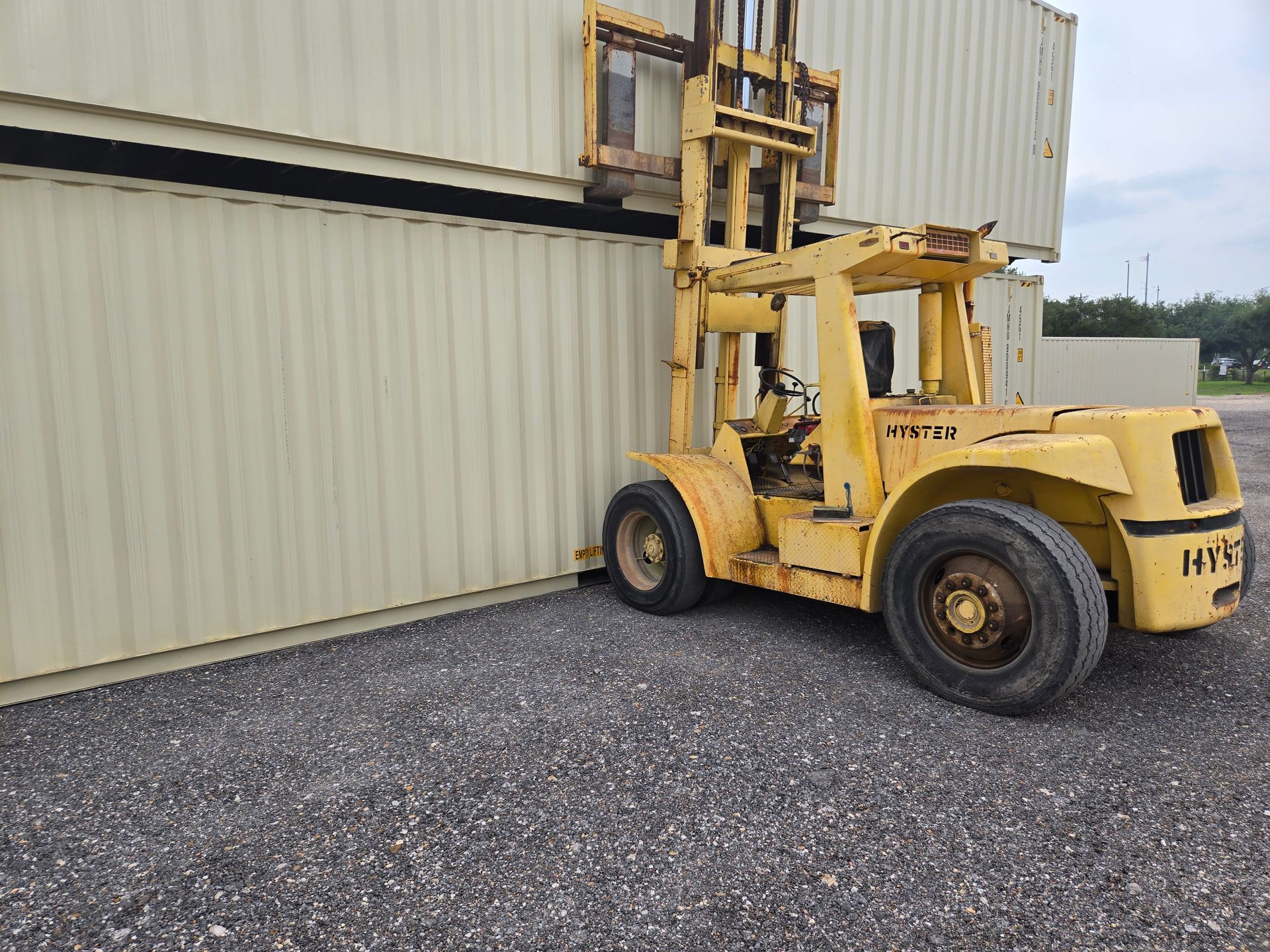 Yellow forklift lifting a shipping container beside a beige metal building on gravel