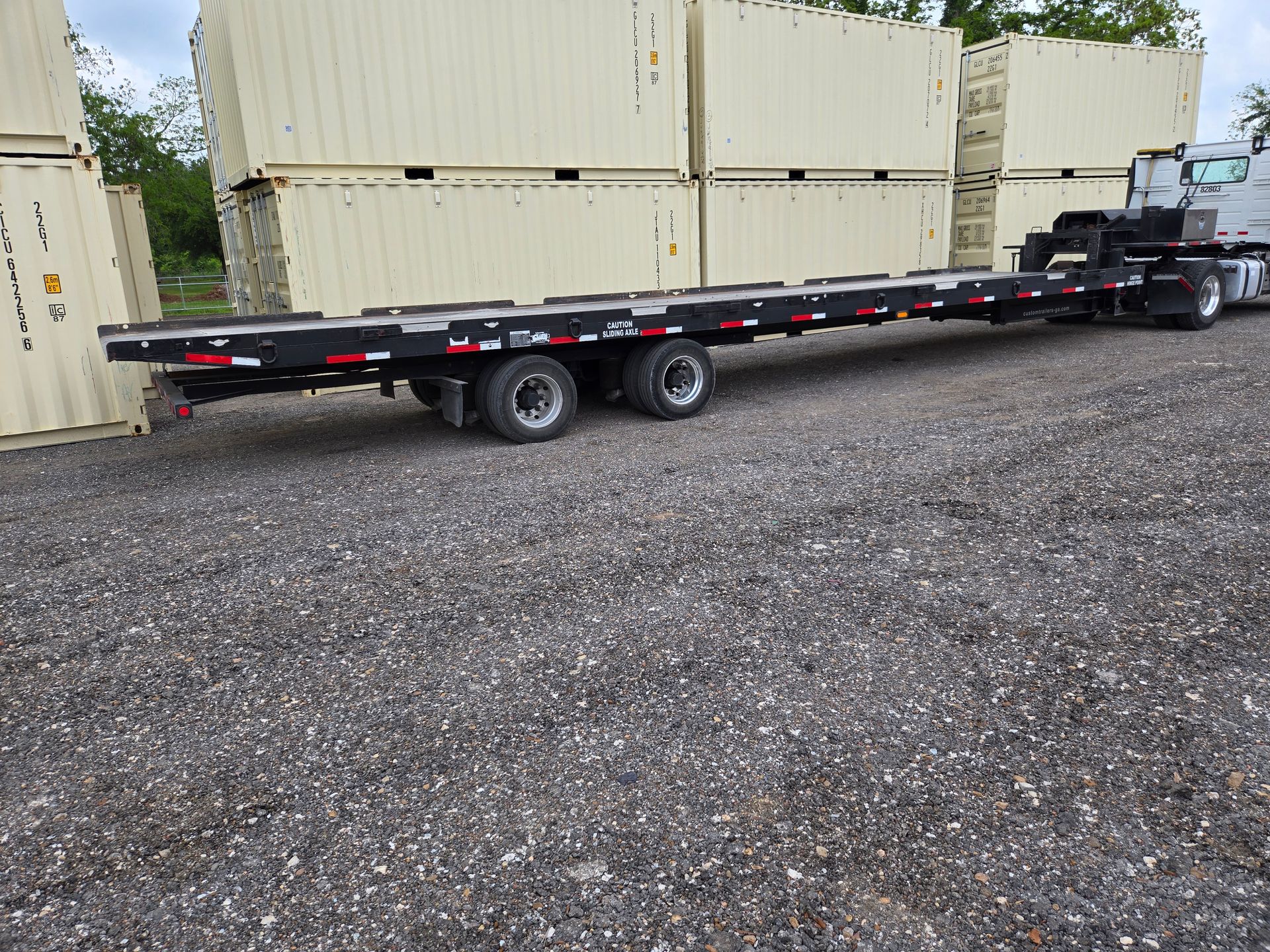 Black flatbed trailer parked on gravel beside stacked beige shipping containers