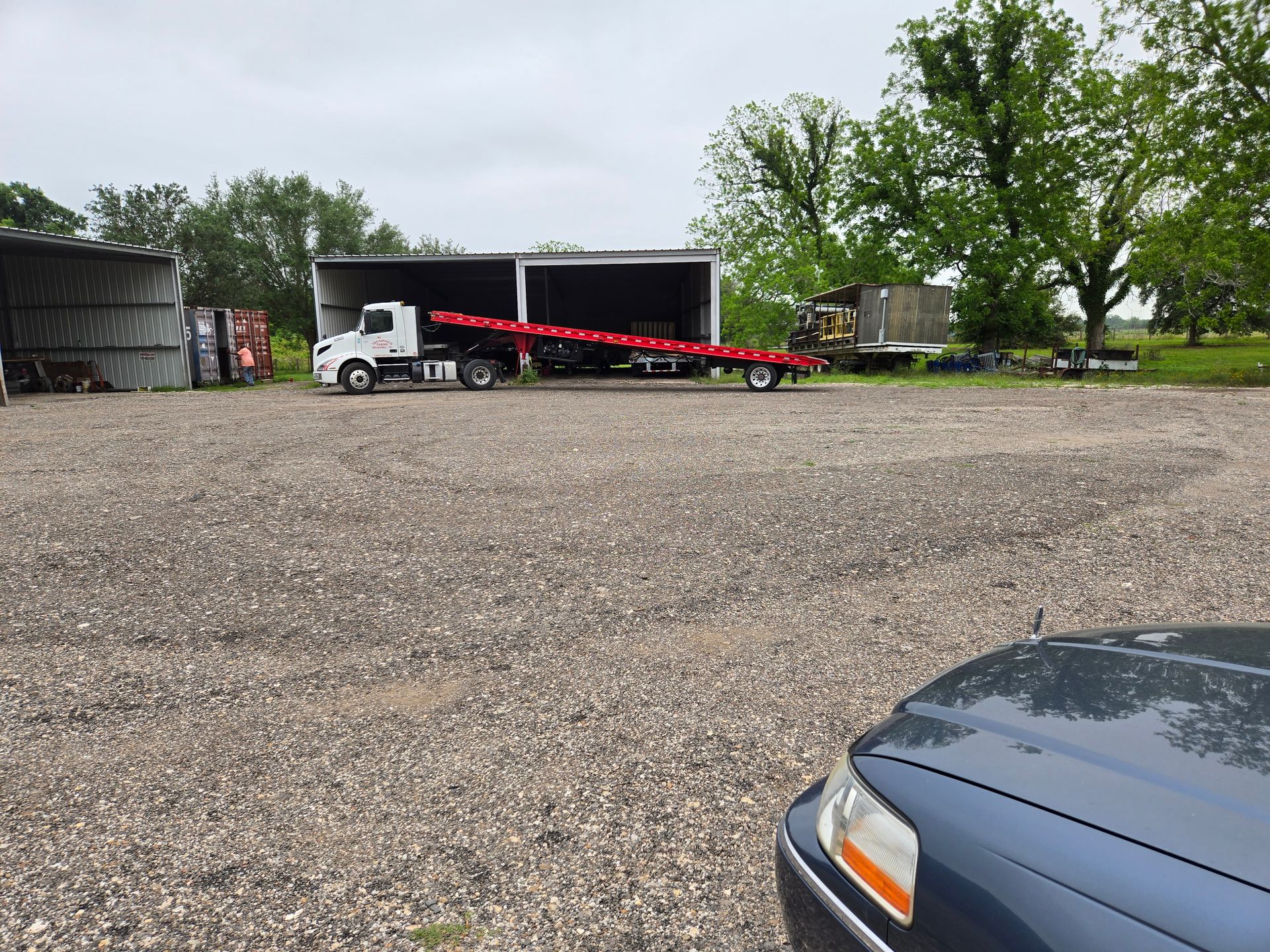 White truck hauling a long red trailer in a gravel lot near open sheds and trees