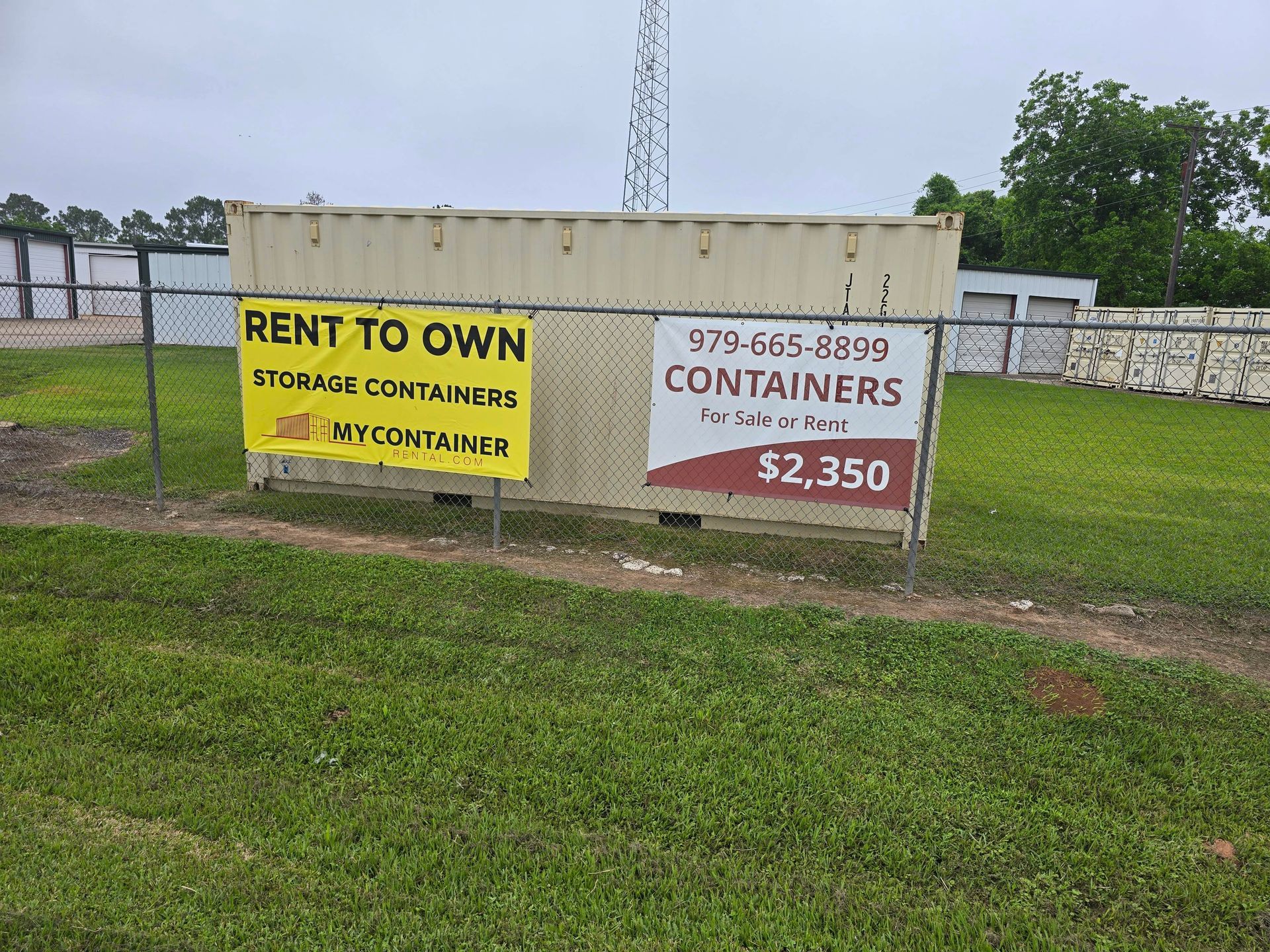 Fence signs advertising rent-to-own storage containers, one yellow and one white, beside a grassy lot