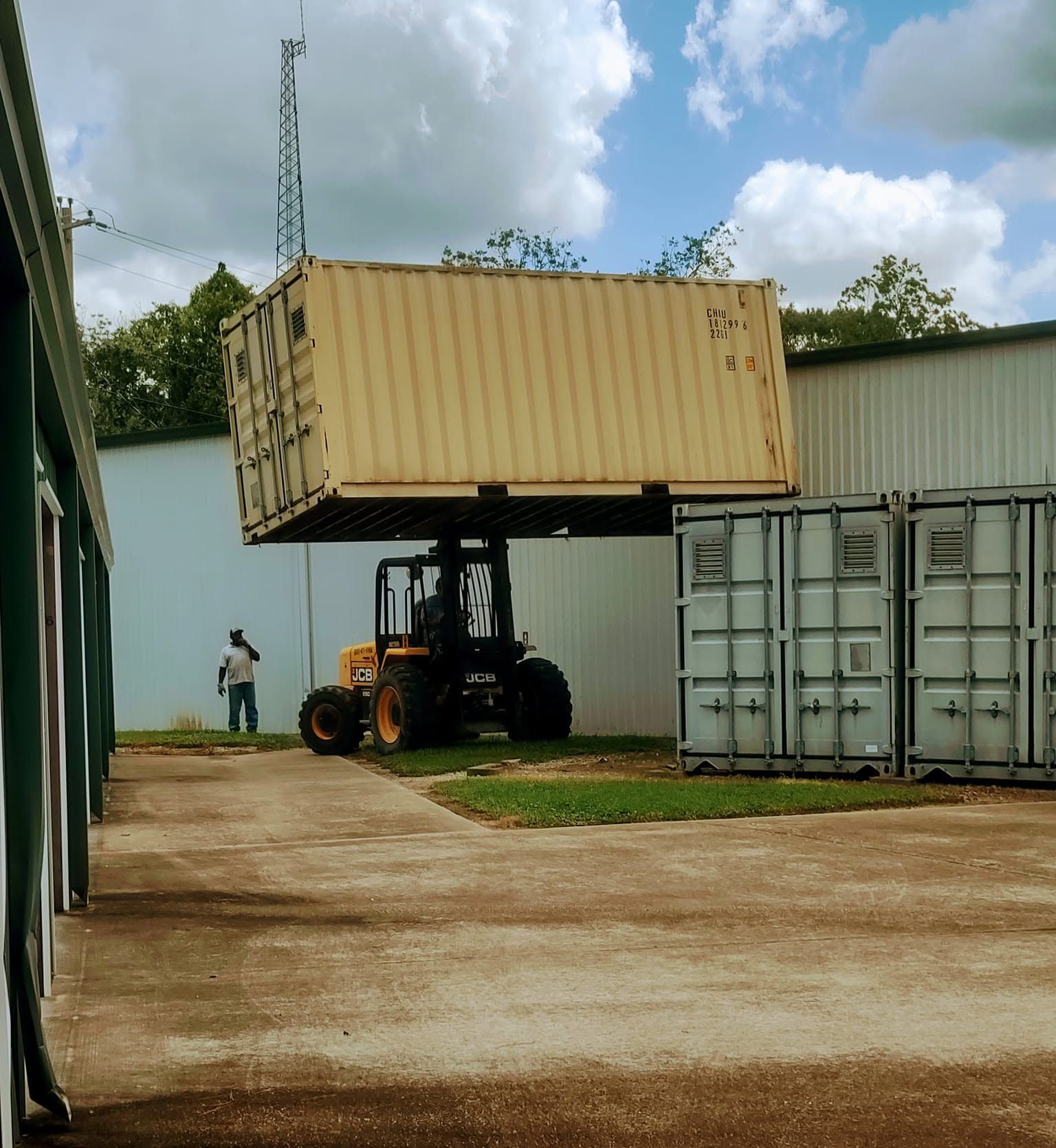 Forklift lifting a beige shipping container beside a warehouse, with another container and a person nearby.