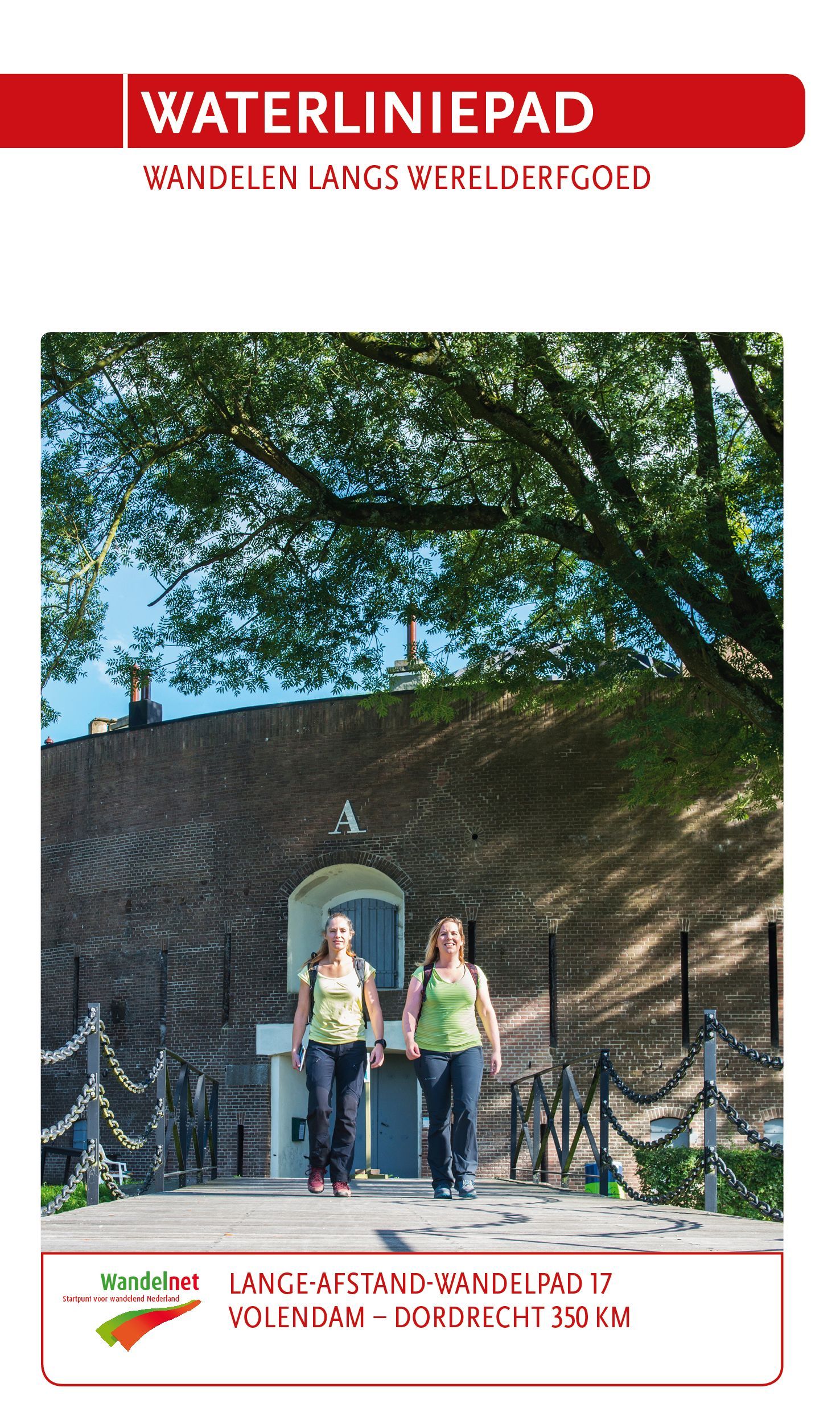 Twee vrouwen lopen over een brug bij een bakstenen gebouw, onder een boom. Titel: "Waterliniepad".