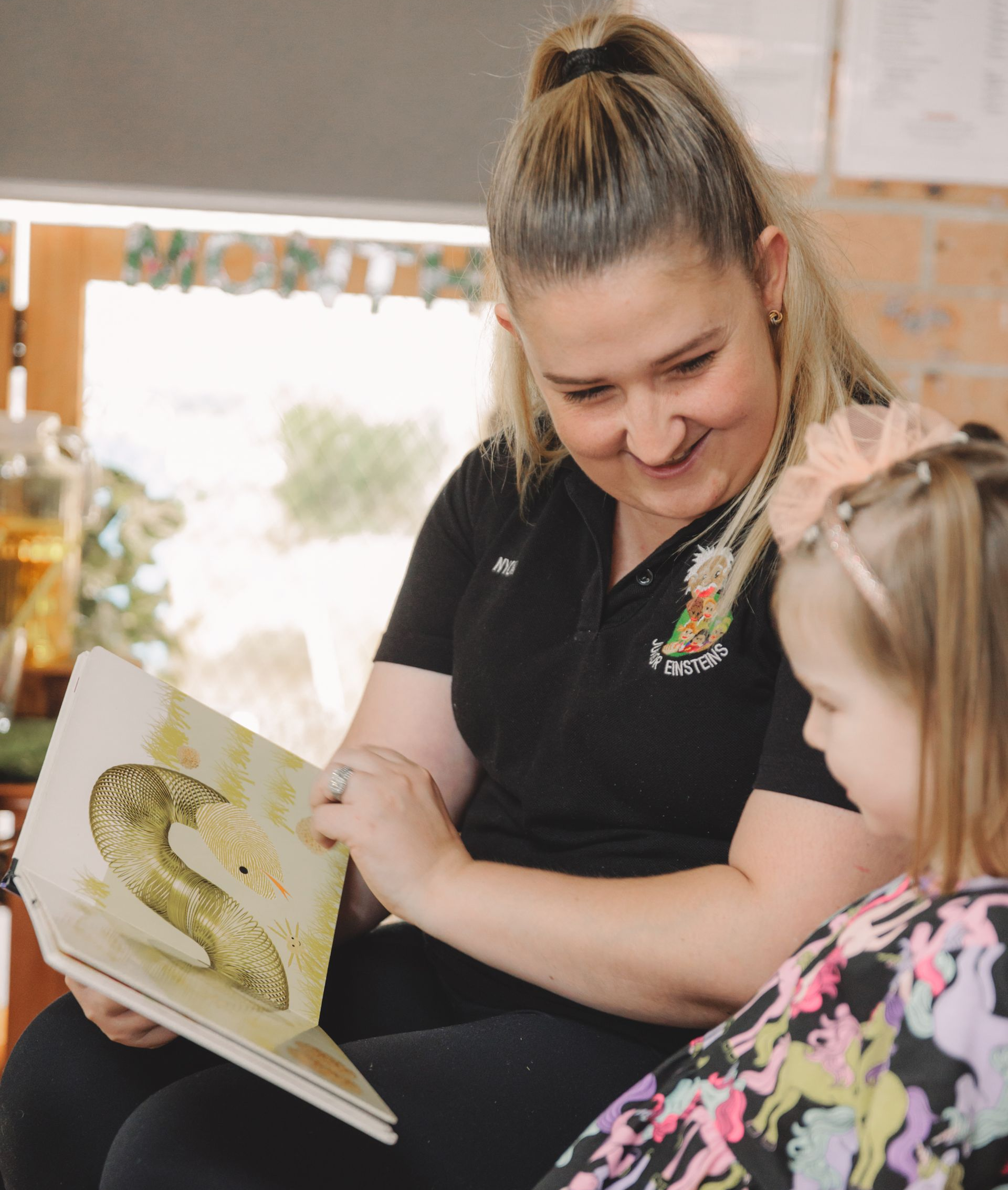 Woman reads to a young child from a book. Both are smiling, in an indoor setting.