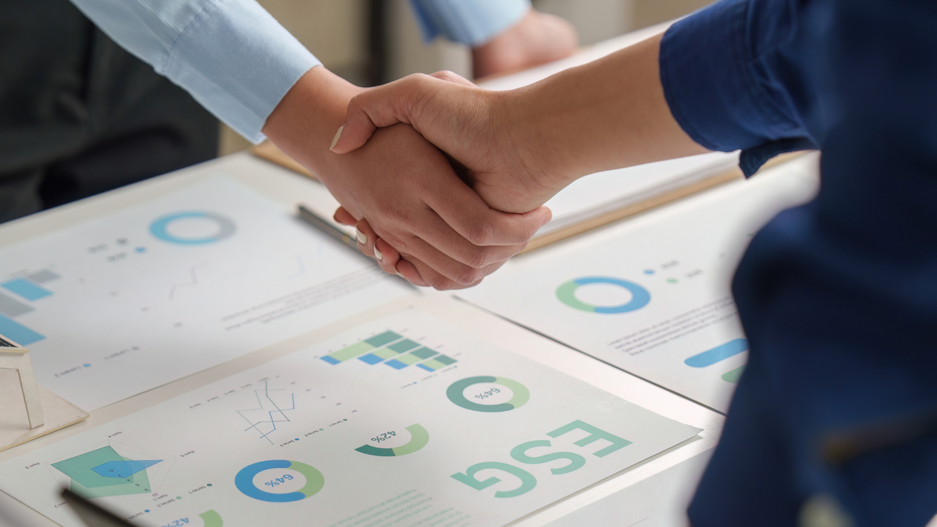 Two people shake hands over printed business charts and graphs on a desk