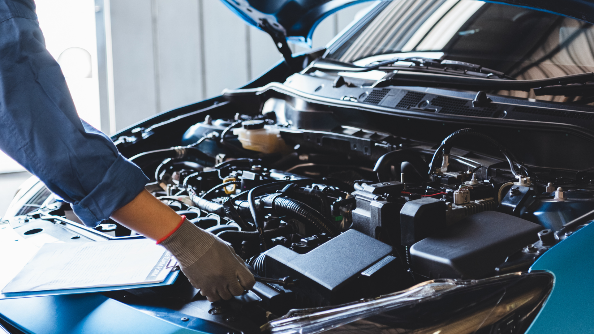 Mechanic inspecting an open car engine in a garage, with a gloved hand over the hood.
