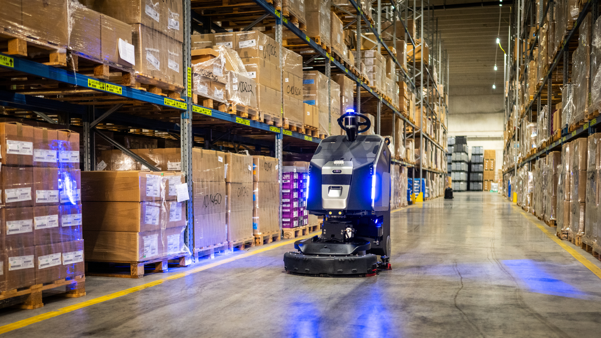 Autonomous warehouse robot driving down an aisle lined with stacked cardboard boxes
