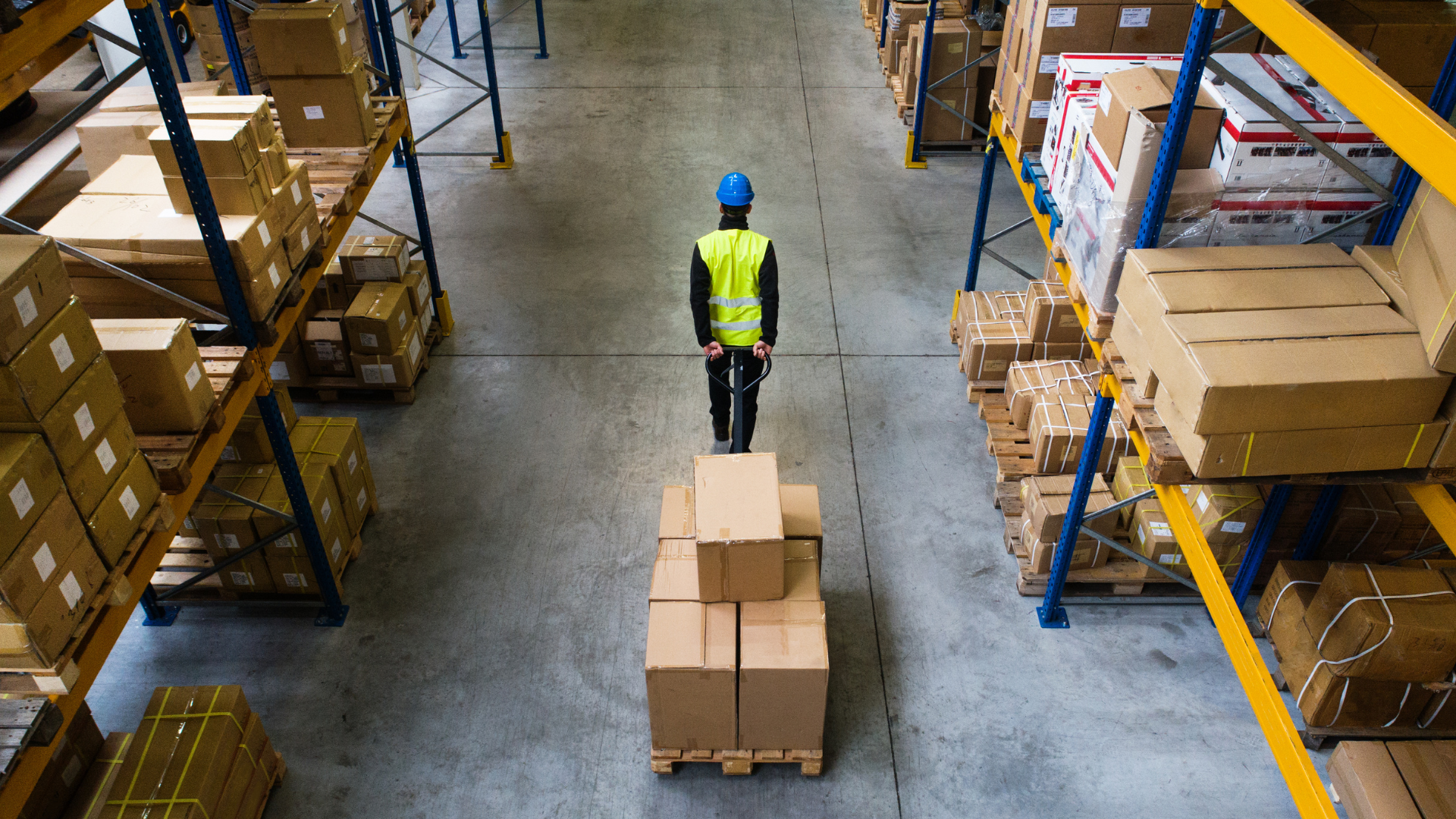Warehouse aisle with a worker in a yellow vest pushing a pallet of boxes between storage racks.