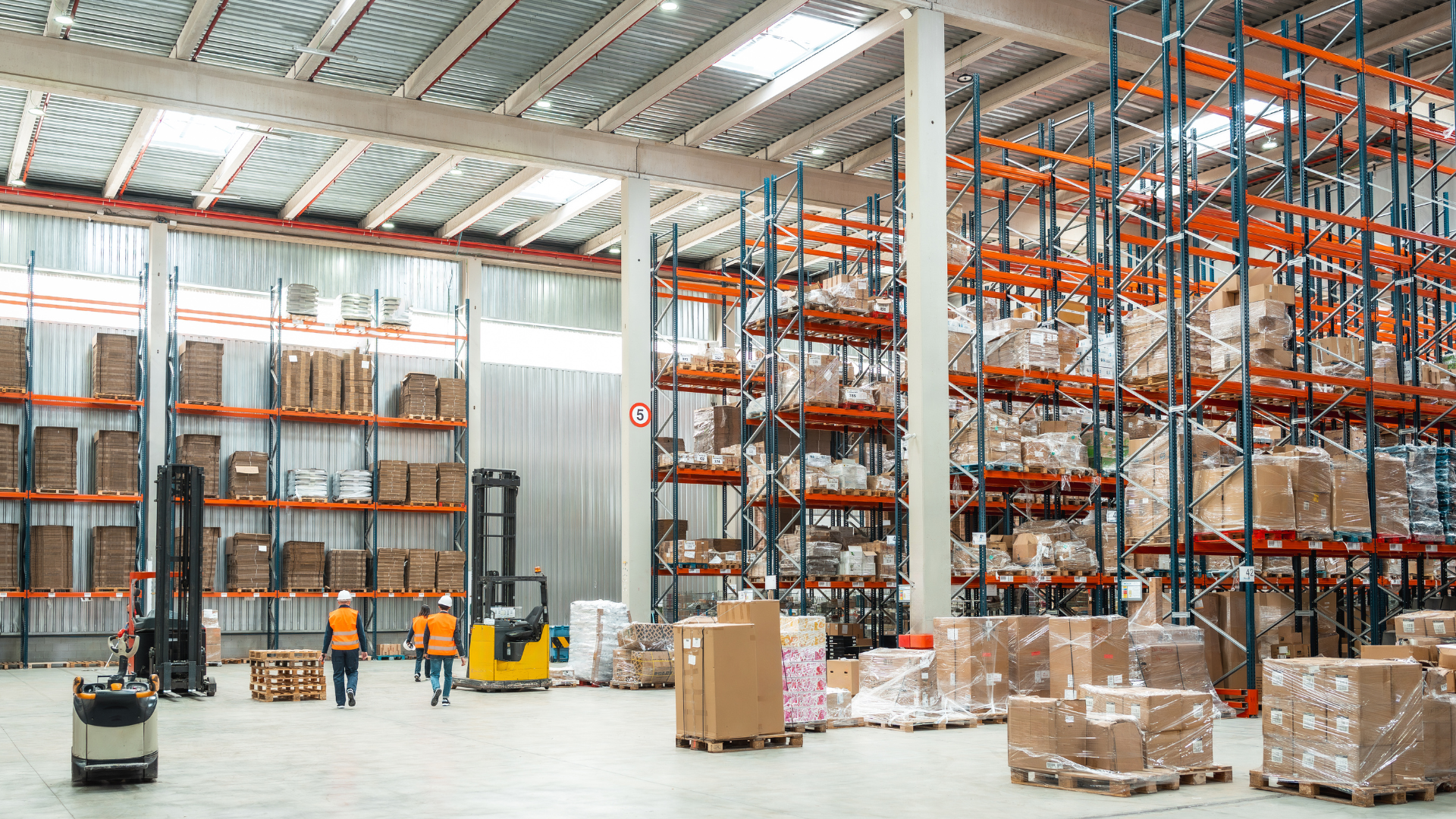 Warehouse with tall orange shelving, stacked pallets, and a forklift, with workers in orange vests nearby