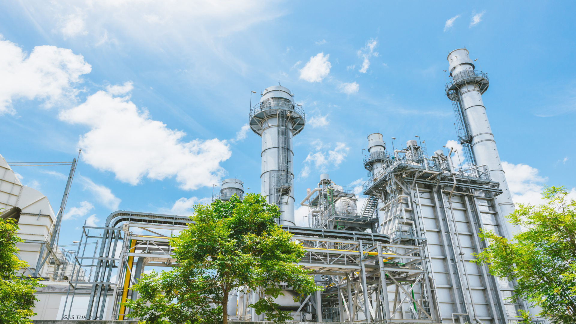 Industrial refinery with tall silver towers and pipes under a blue sky, framed by green trees