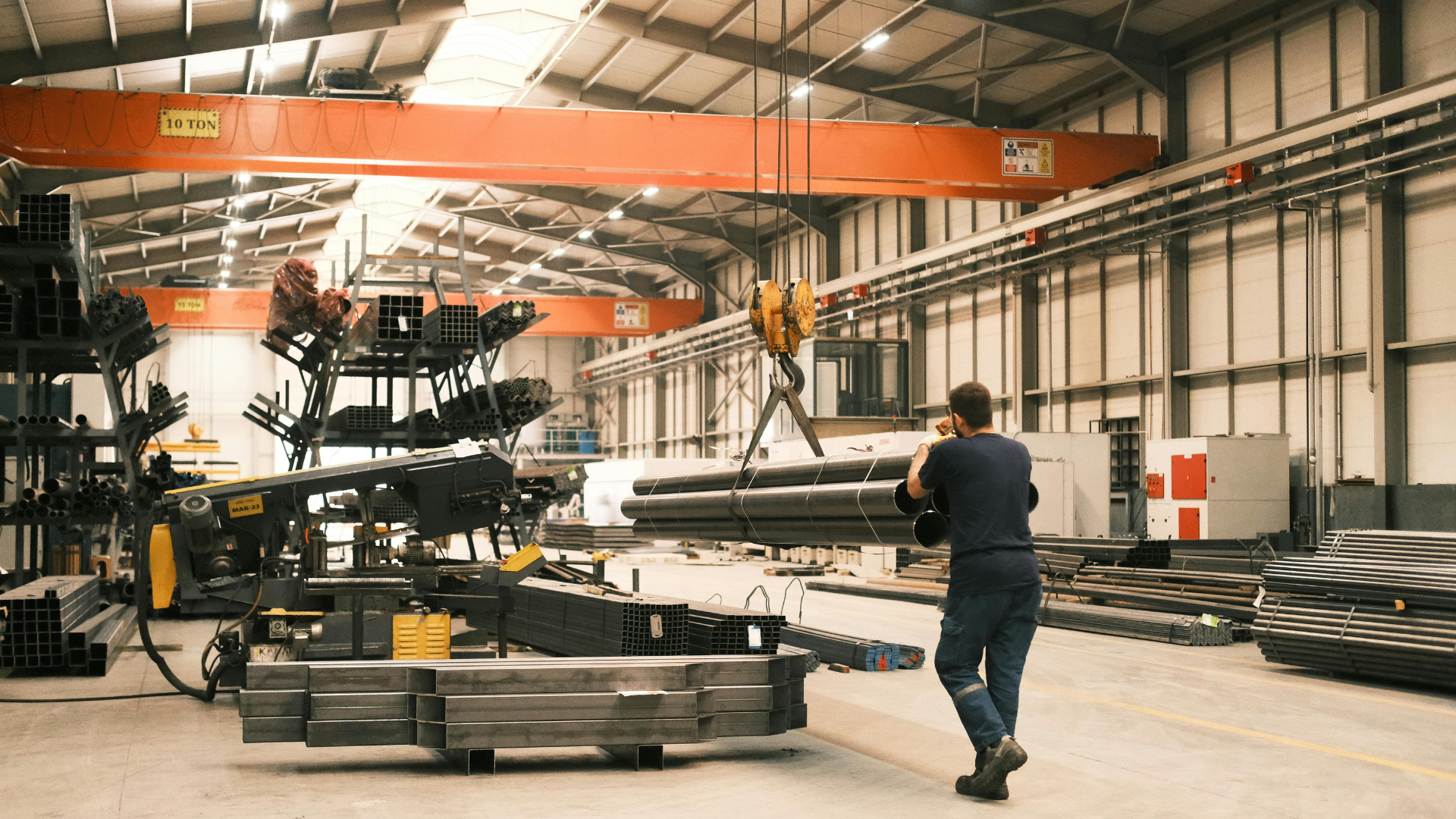 Worker in a warehouse moving long metal beams near stacked materials and a forklift