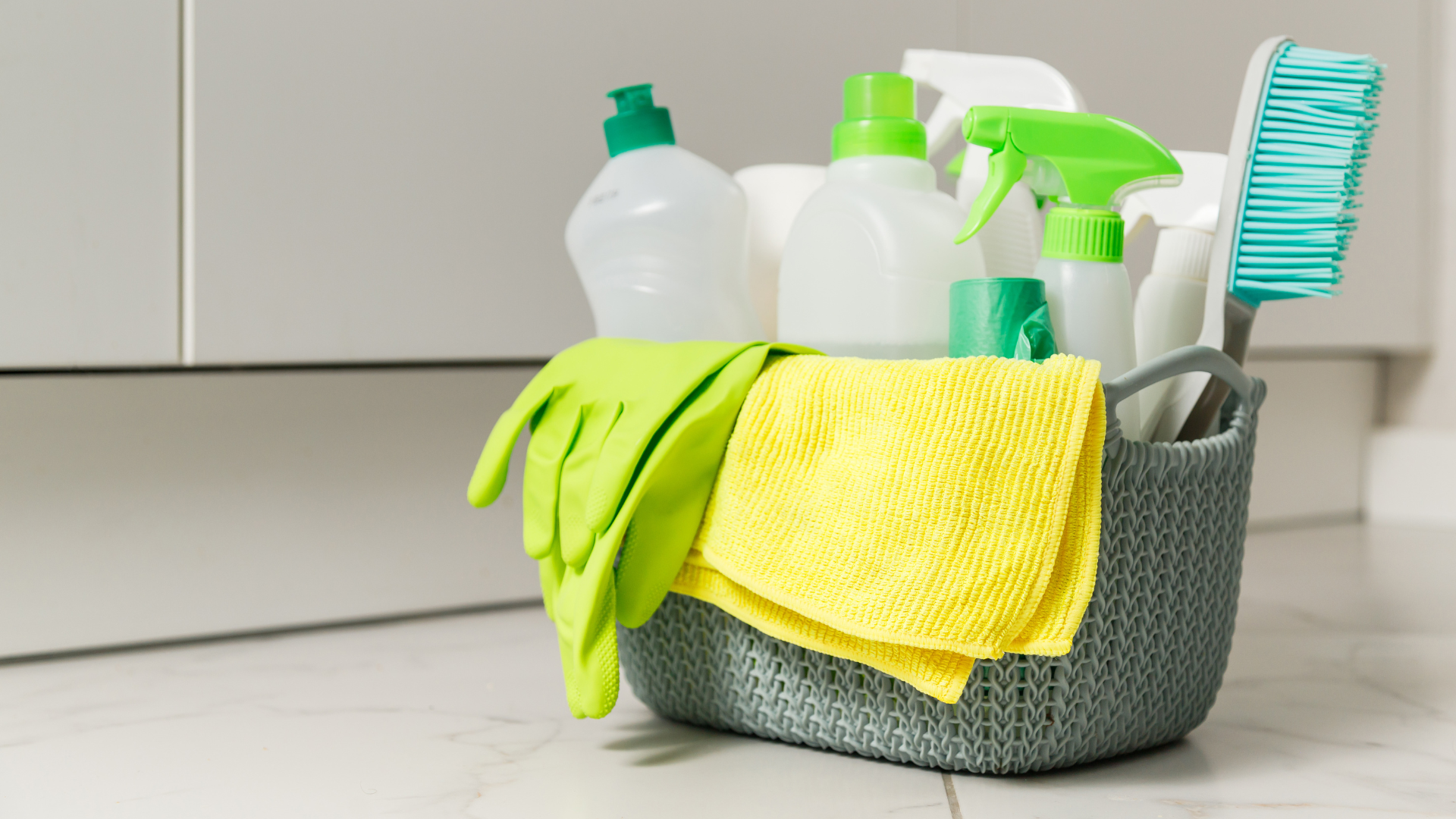 Cleaning supplies in a gray basket, with yellow and green cloths and spray bottles on a counter.