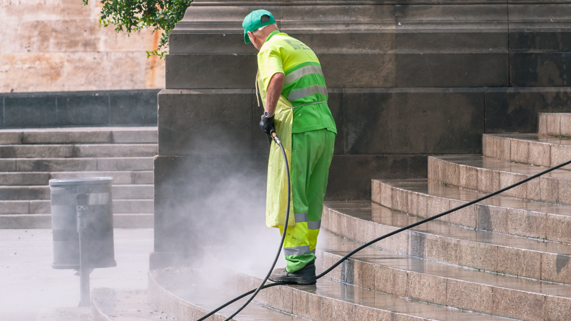 Worker in green safety gear pressure-washing stone steps outdoors.
