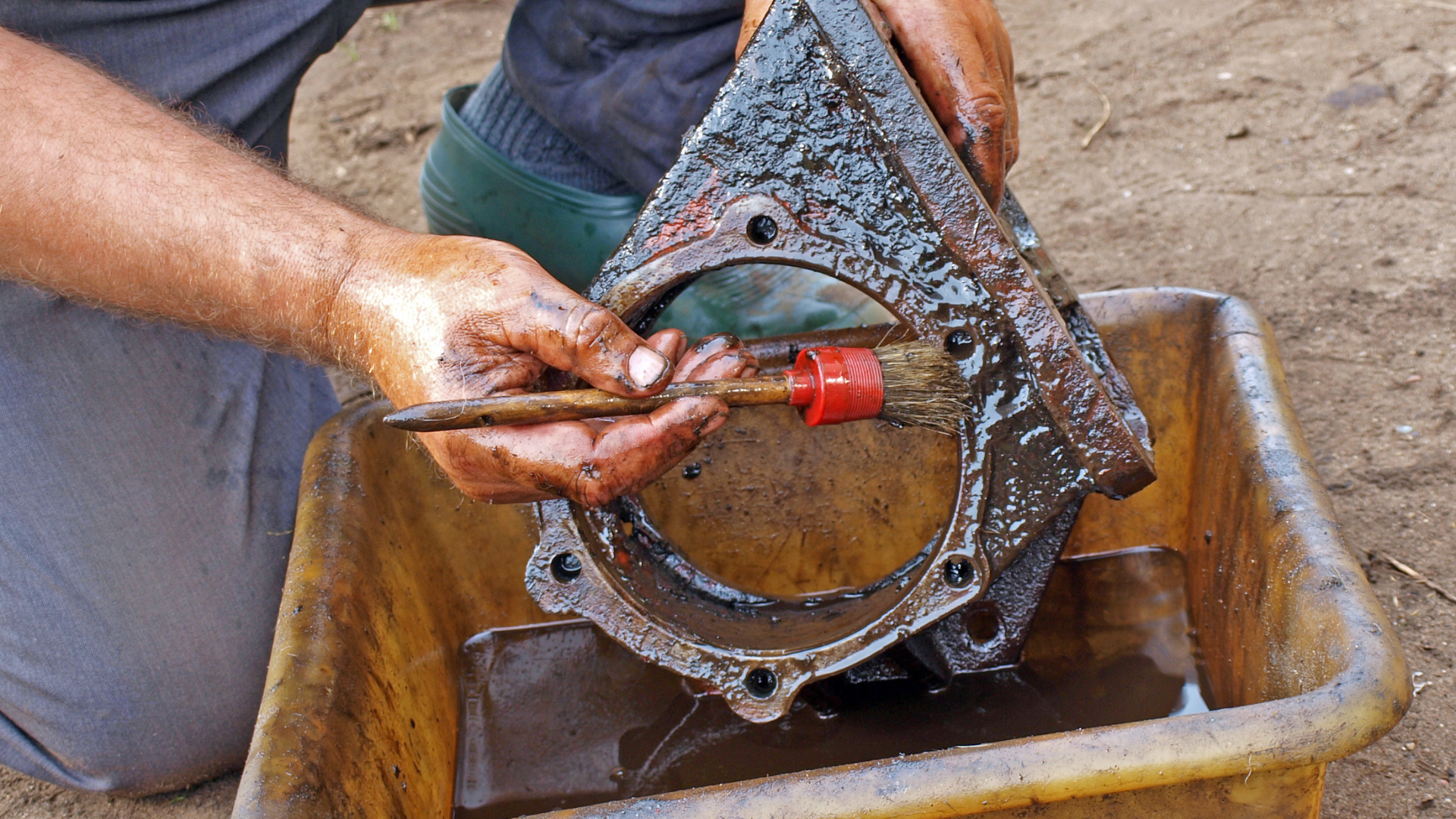 Hands cleaning a rusty metal part with a brush over a basin of water