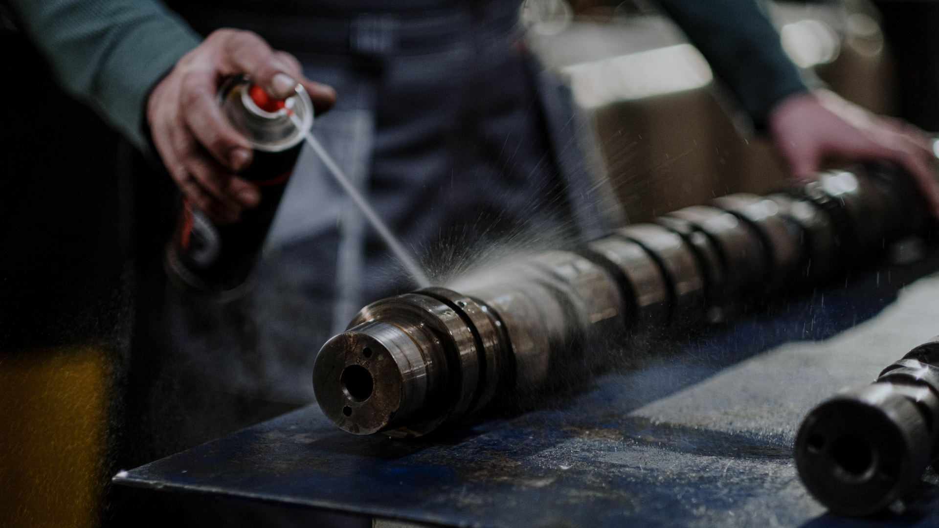 Person sprays lubricant onto a metal shaft on a workbench.