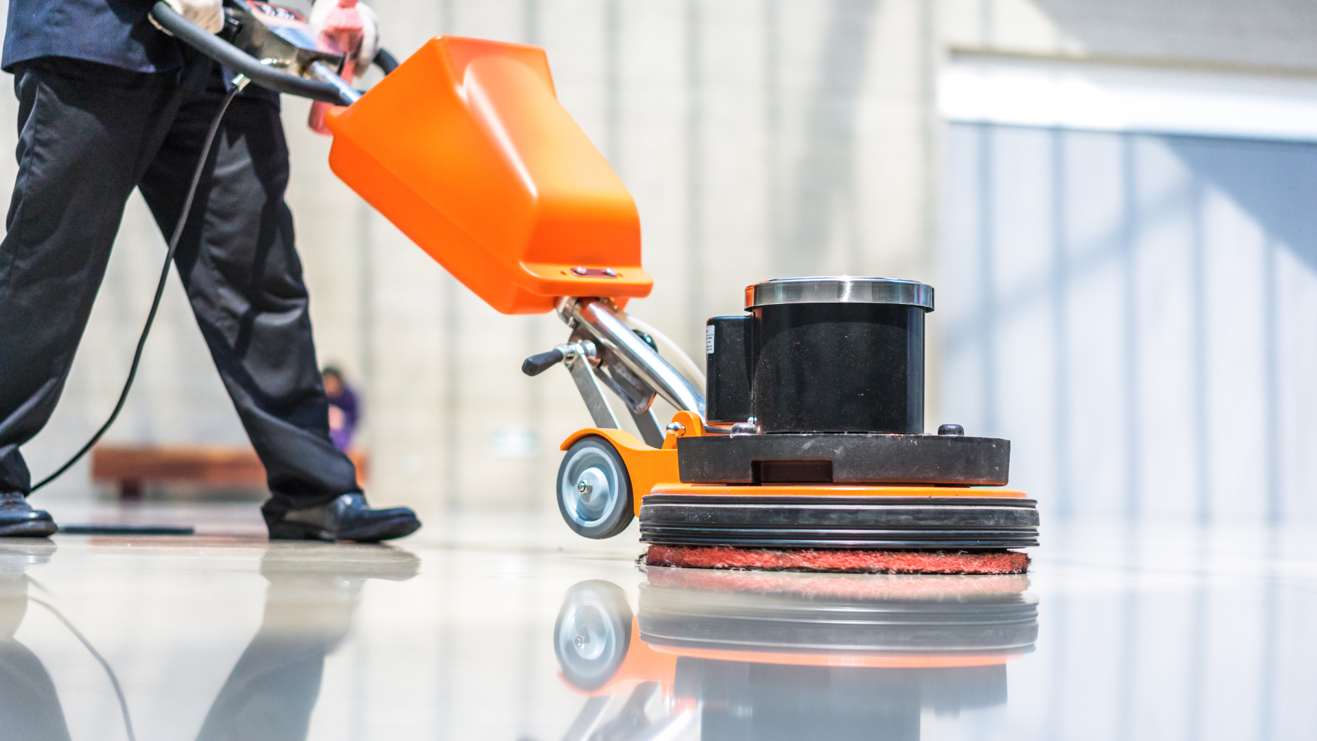 Person operating an orange floor-cleaning machine on a polished indoor floor.