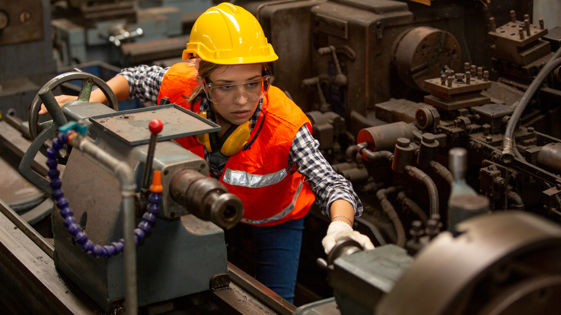 Worker in yellow hard hat and orange safety vest inspecting machinery in an industrial workshop