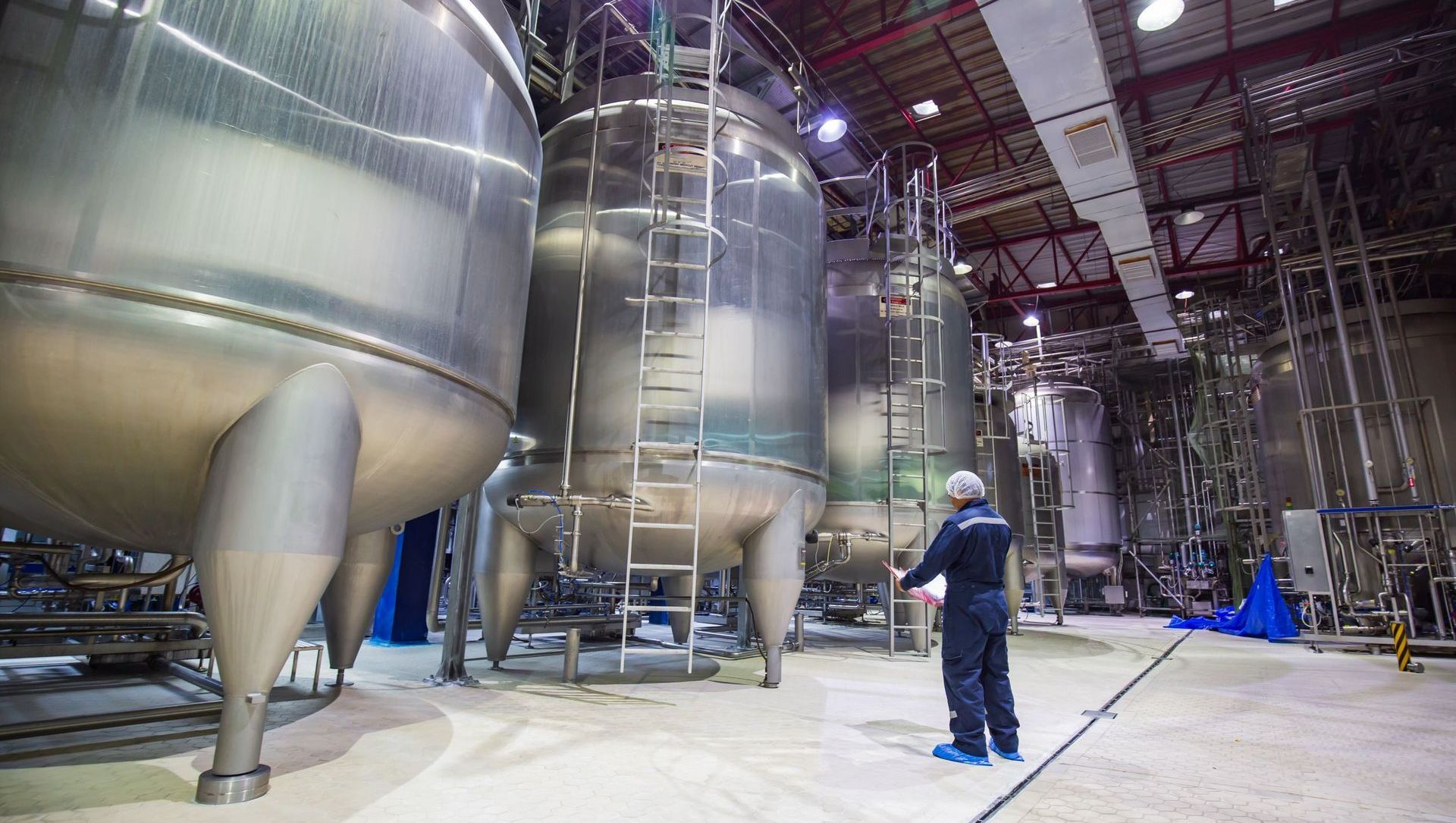 A man is standing in front of a row of stainless steel tanks in a factory.
