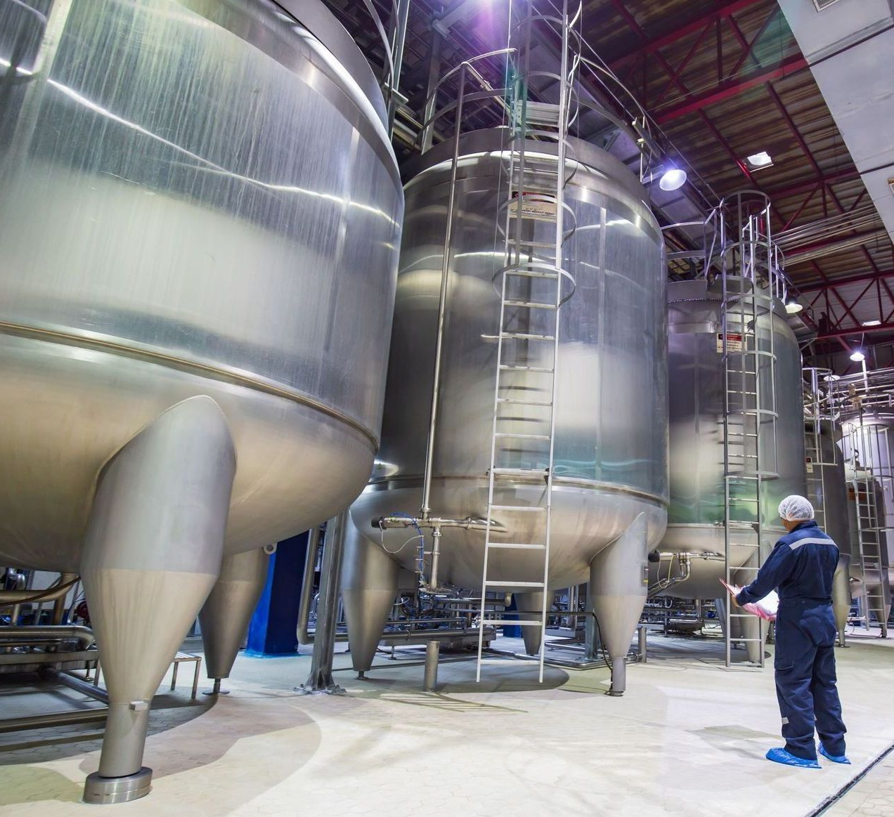 Worker in blue overalls inspecting large stainless steel tanks in a factory setting.