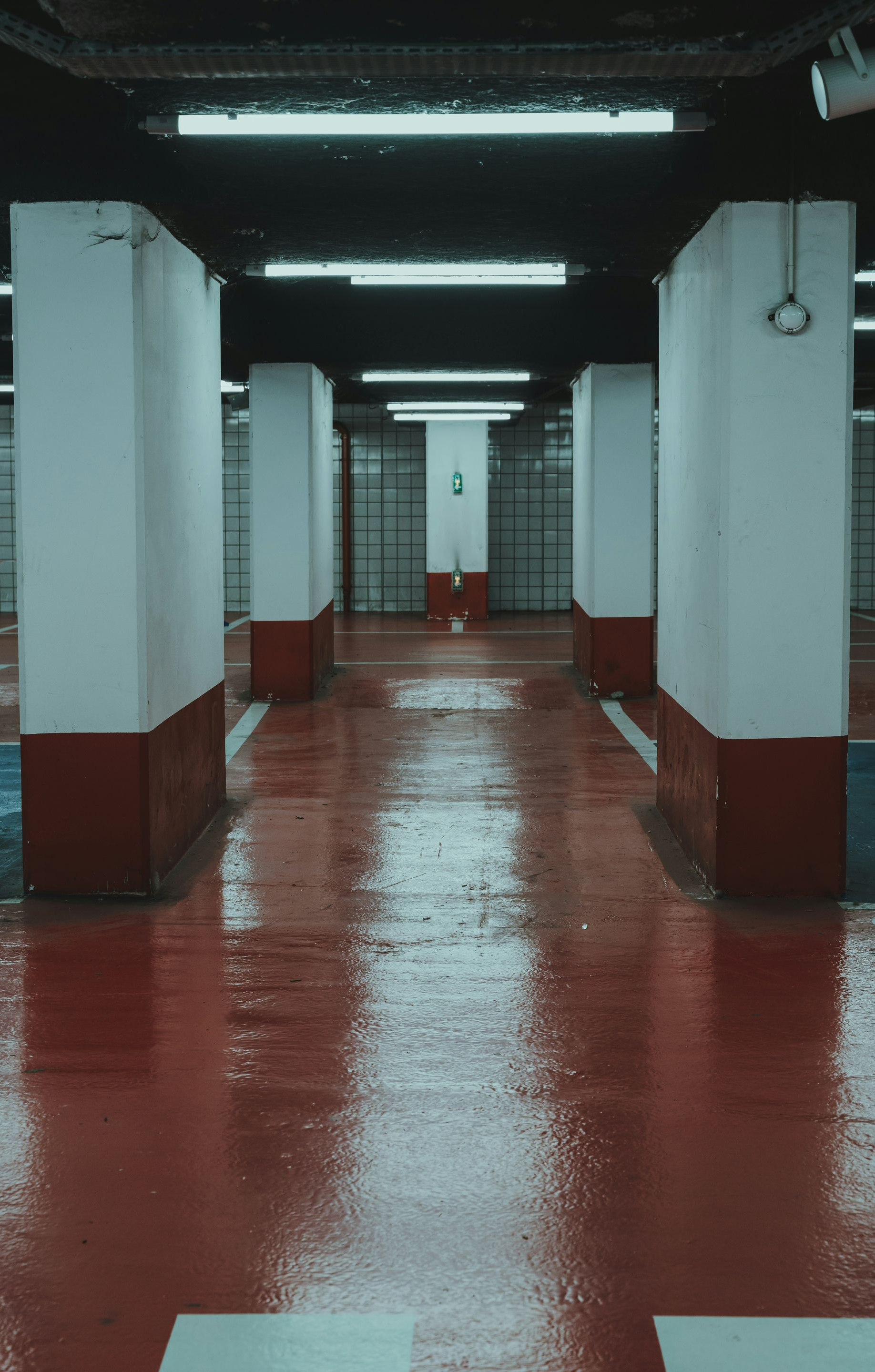 Empty parking garage, red floor reflecting lights, white pillars with red bases, somber atmosphere.