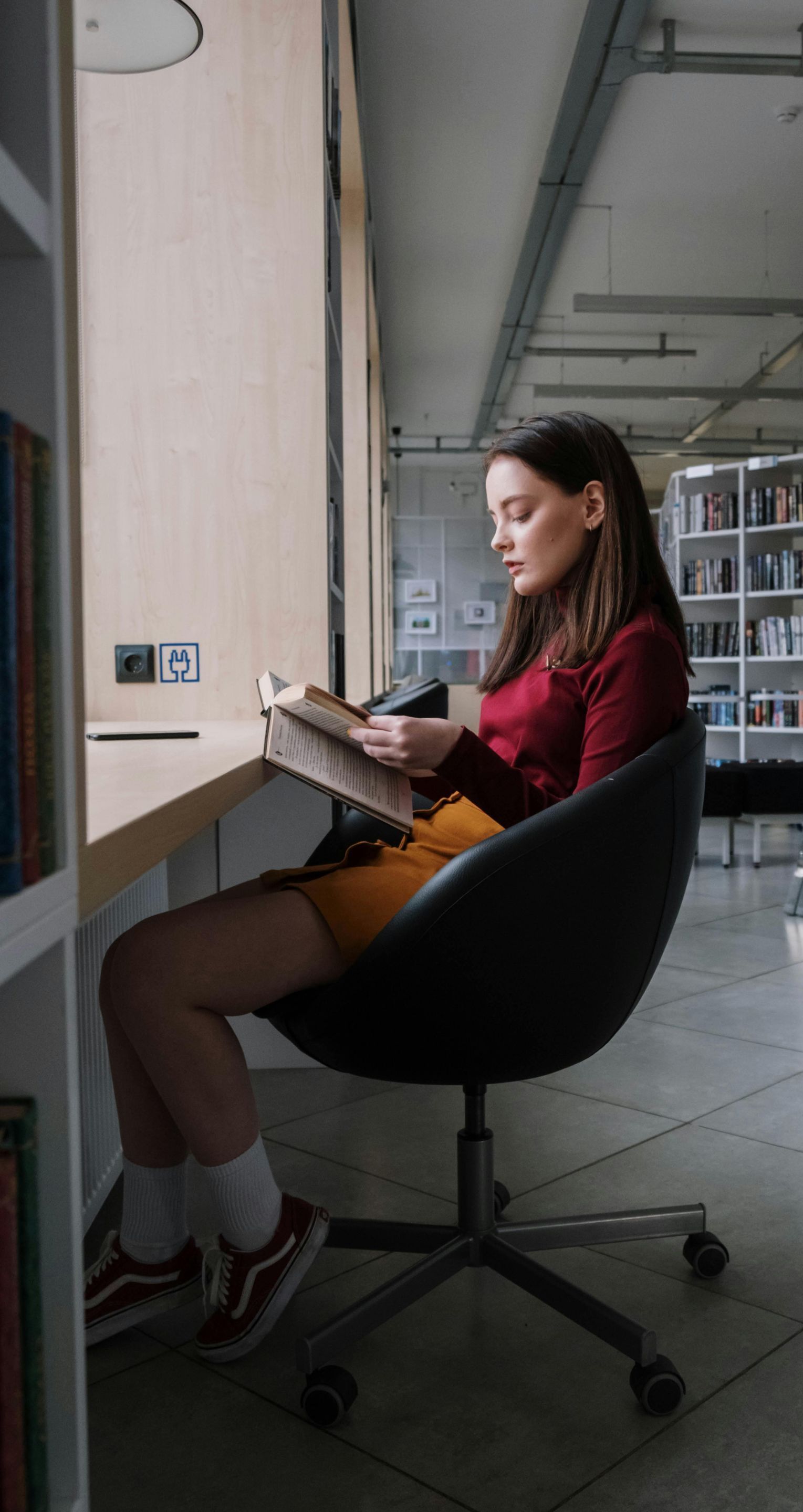 Woman reading a book in a library, seated in a modern chair by a window. She's wearing a red shirt and yellow skirt.