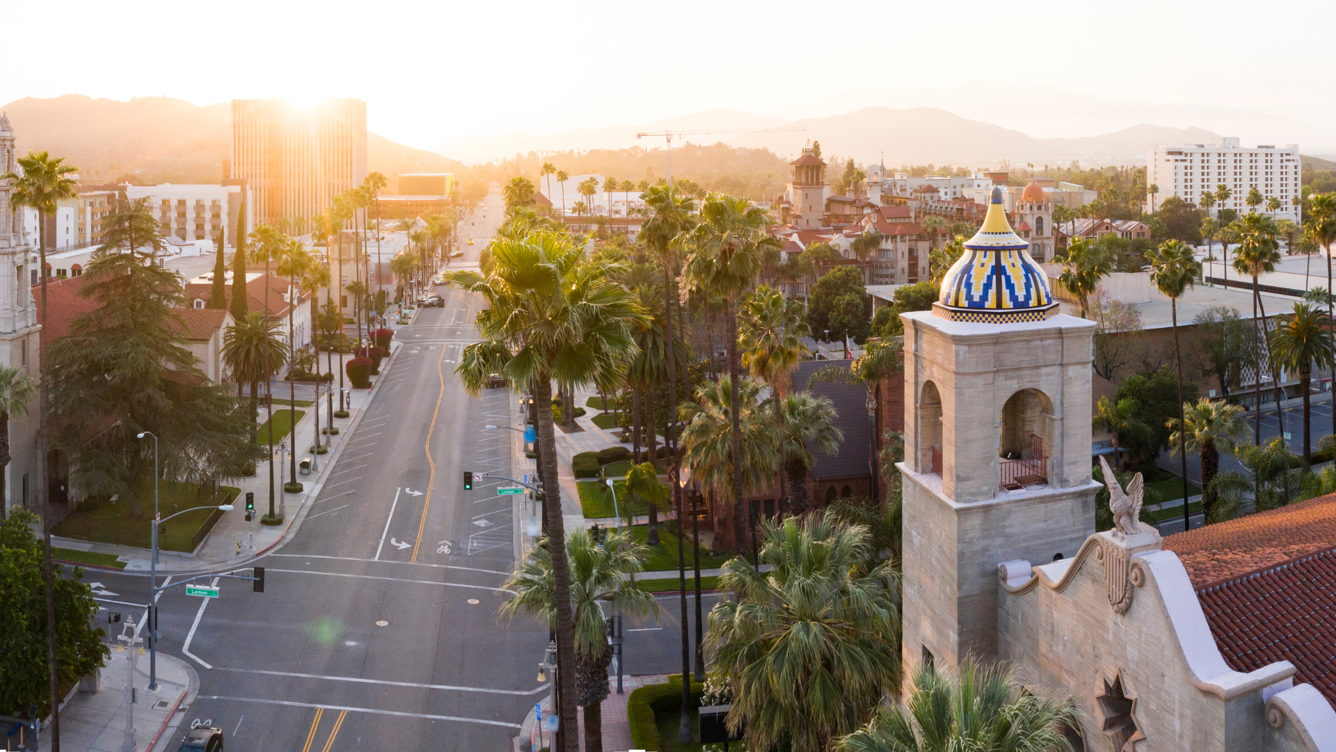 City street lined with palm trees, buildings, and a church tower with a blue and white dome. Sun setting in the distance.