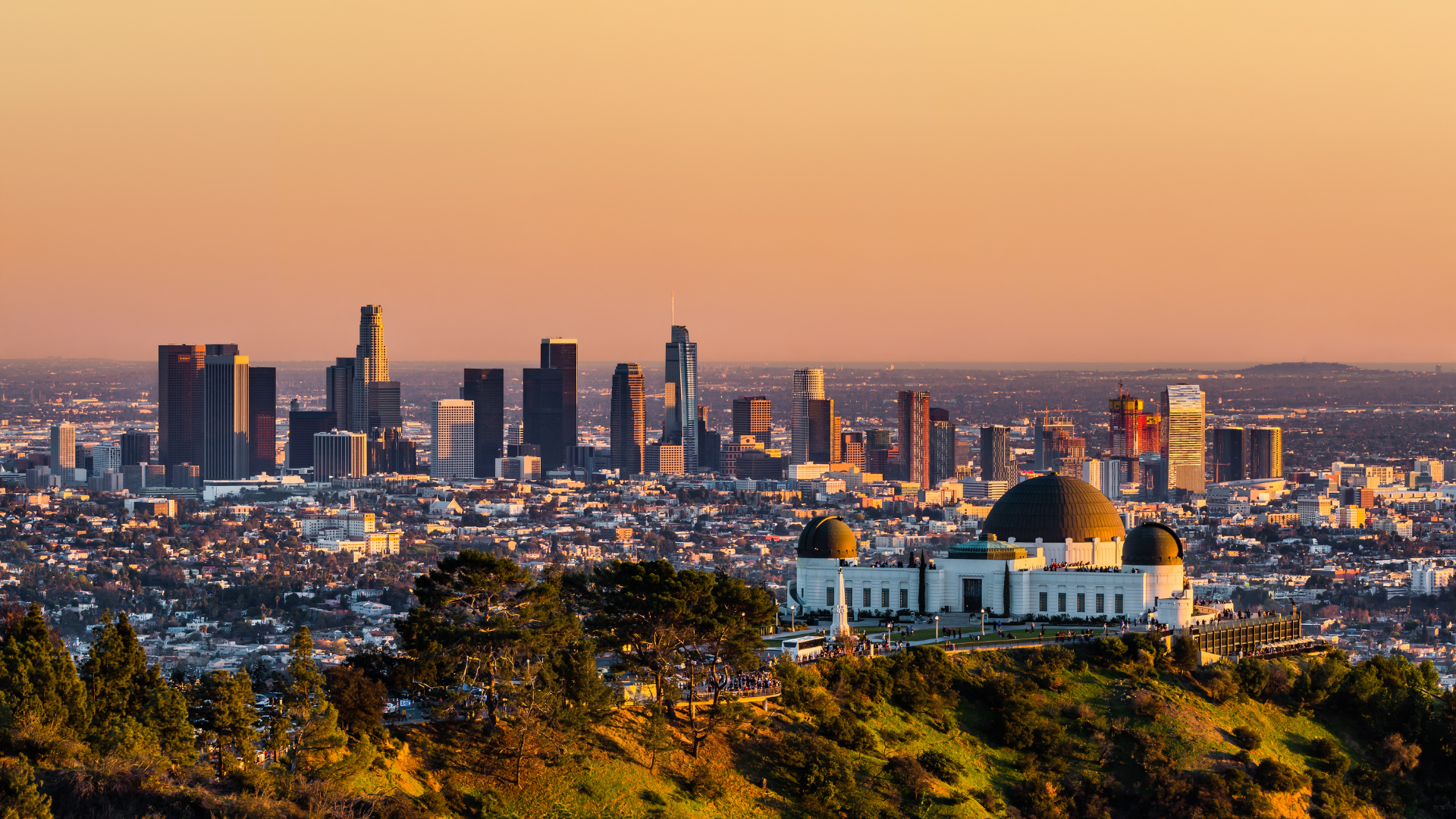 Los Angeles skyline at sunset, with the Griffith Observatory in the foreground, against an orange sky.