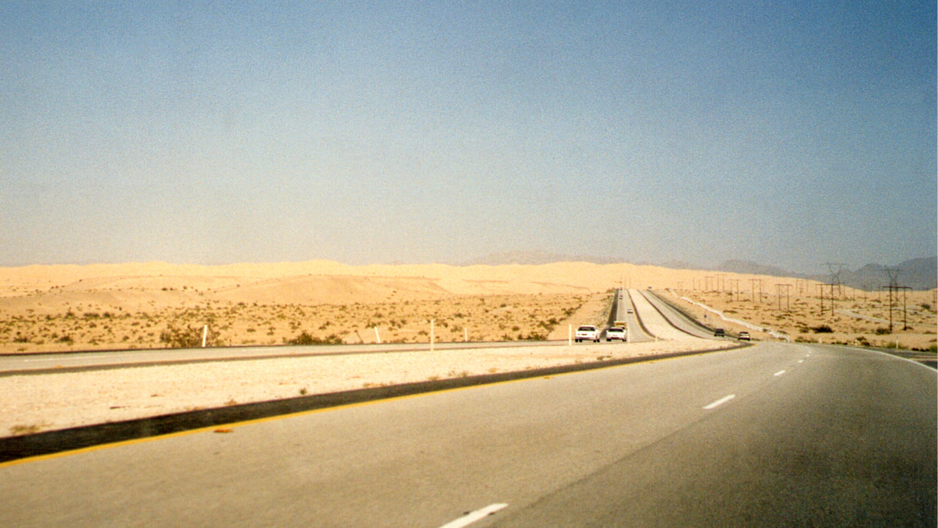 A desert highway with two vehicles, sandy hills, and a clear blue sky.