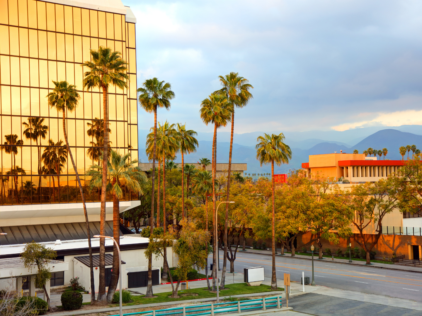 Palm trees in front of golden glass building reflect sunset. Mountains and cloudy sky in background.