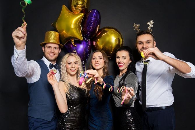 Five people at a party in hats and ties, holding confetti poppers and balloons against a dark backdrop