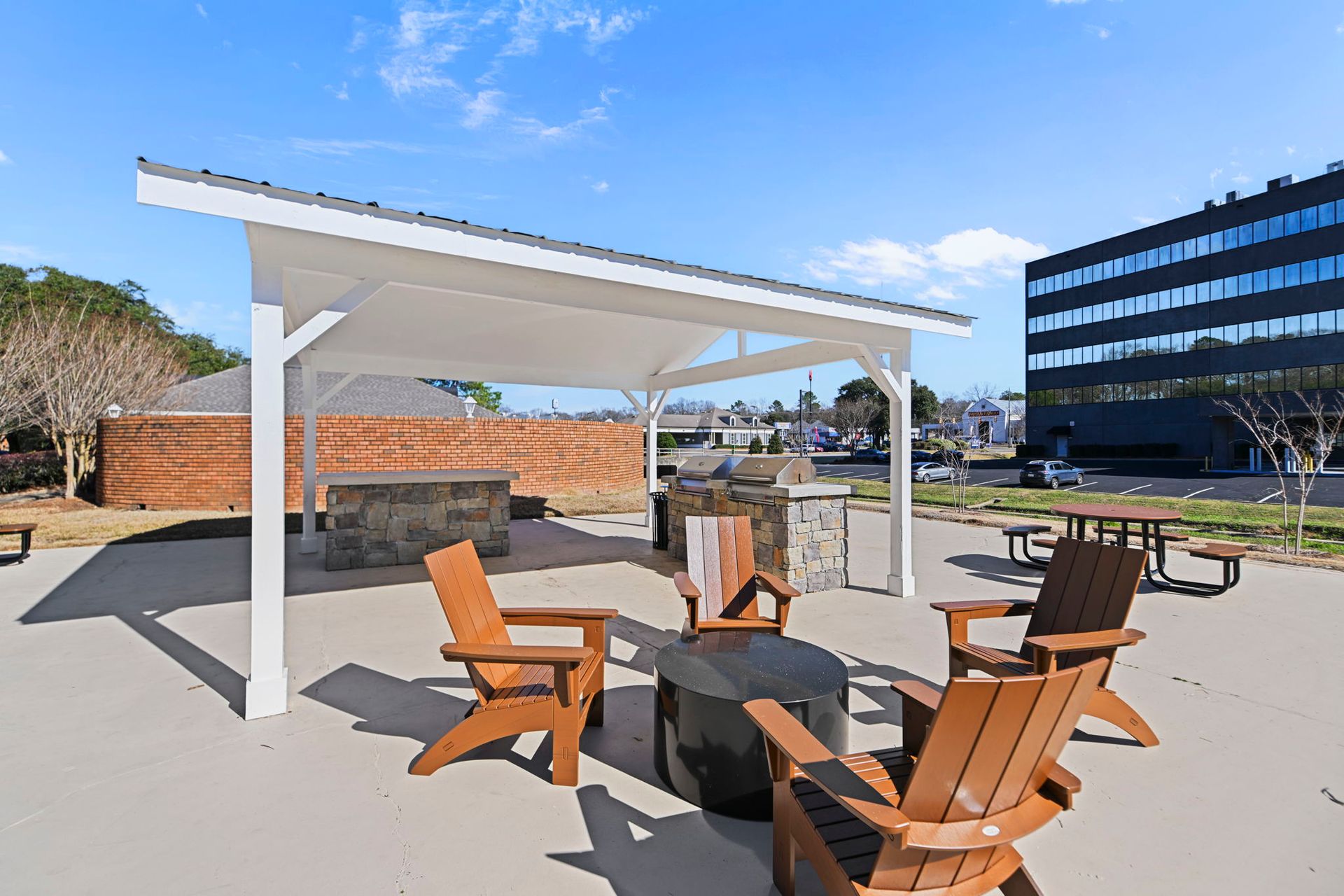 Outdoor patio with a covered pergola, stone grill station, fire pit, and several brown chairs under a clear blue sky.