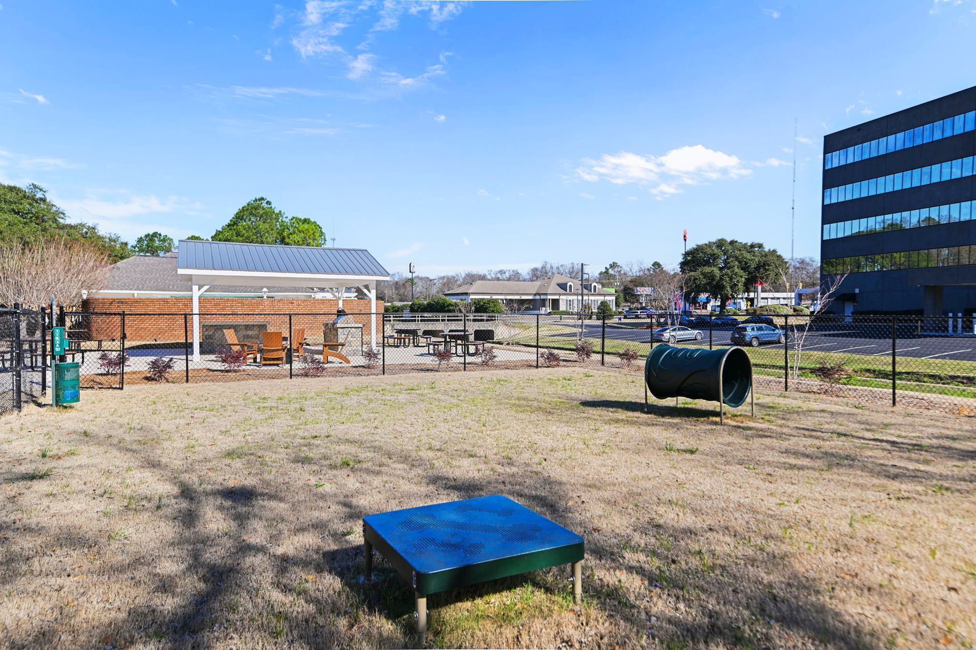 An outdoor dog park with a play tunnel, a square platform, and a covered patio area under a bright blue sky.