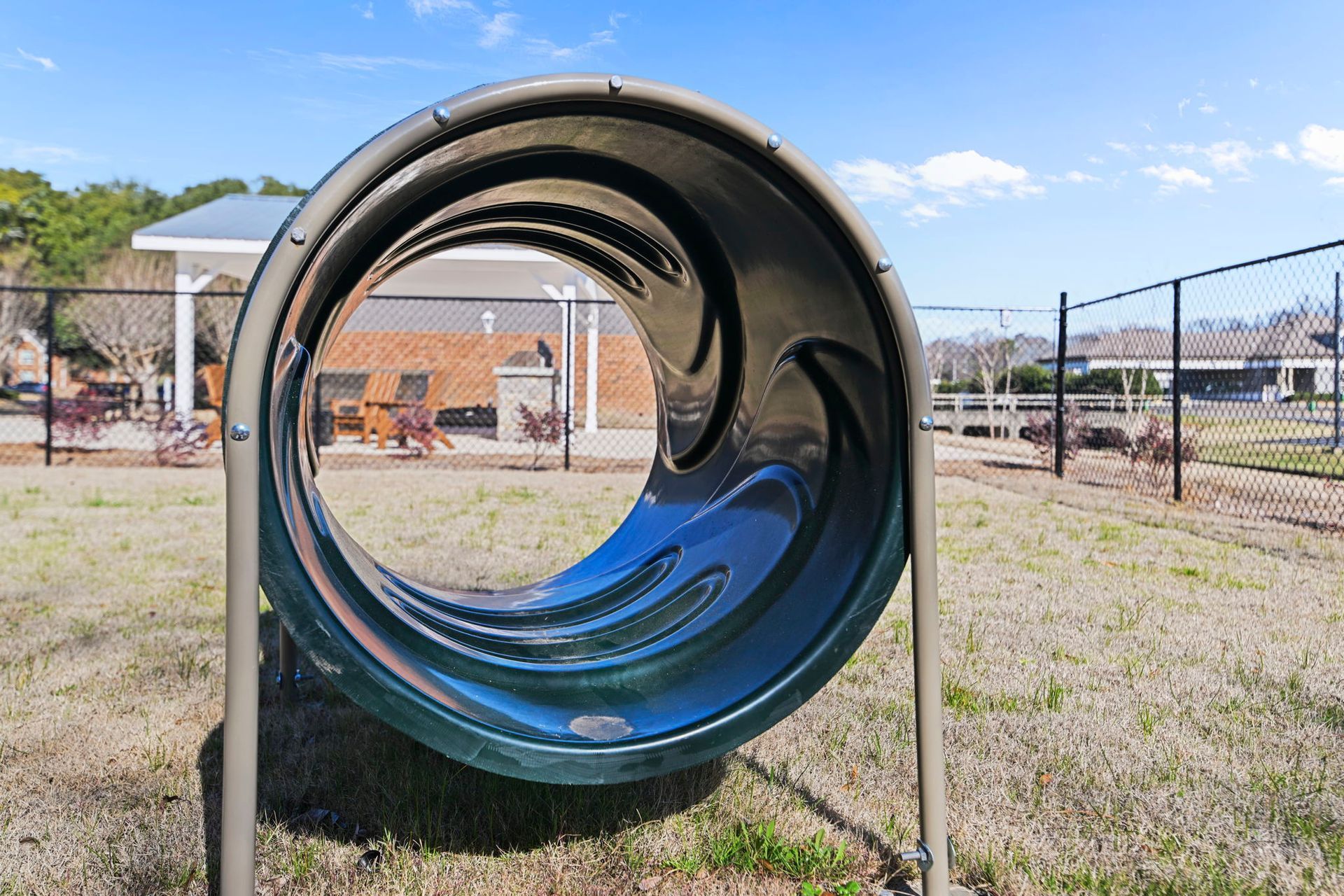 A green dog agility tunnel sits on a dry, grassy field in a fenced dog park under a clear blue sky.