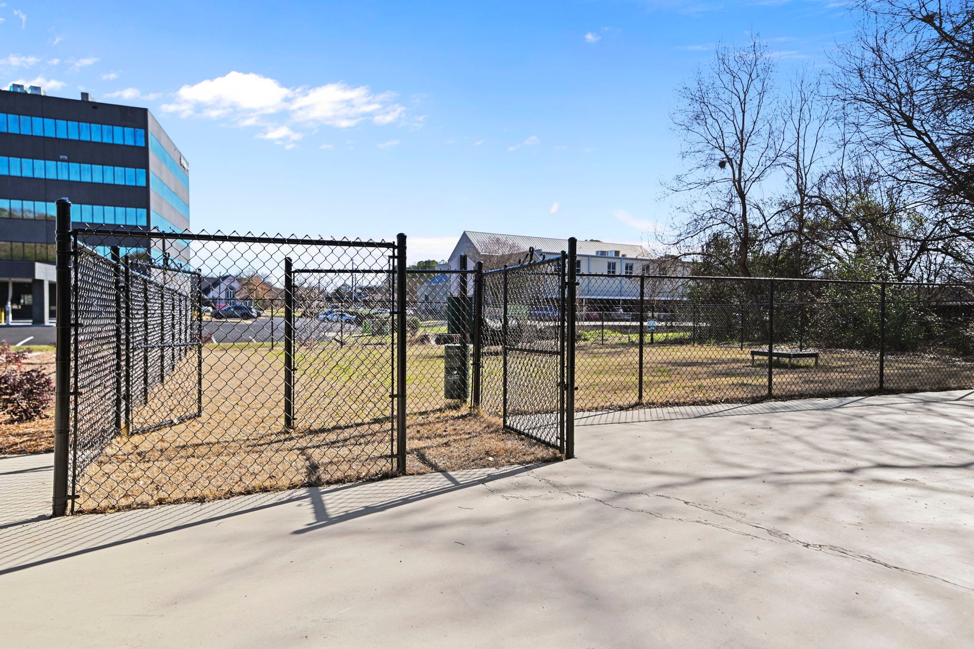 A chain-link fenced enclosure sits on a concrete lot next to a large glass office building under a blue sky.