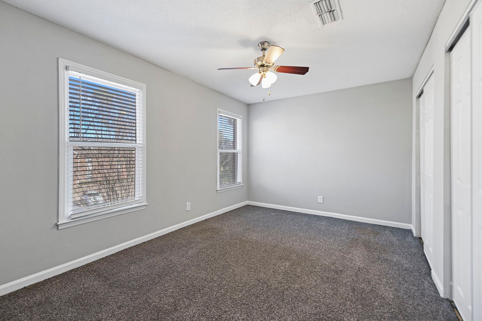 Empty room with gray walls, dark carpet, two windows with blinds, a ceiling fan, and white doors.